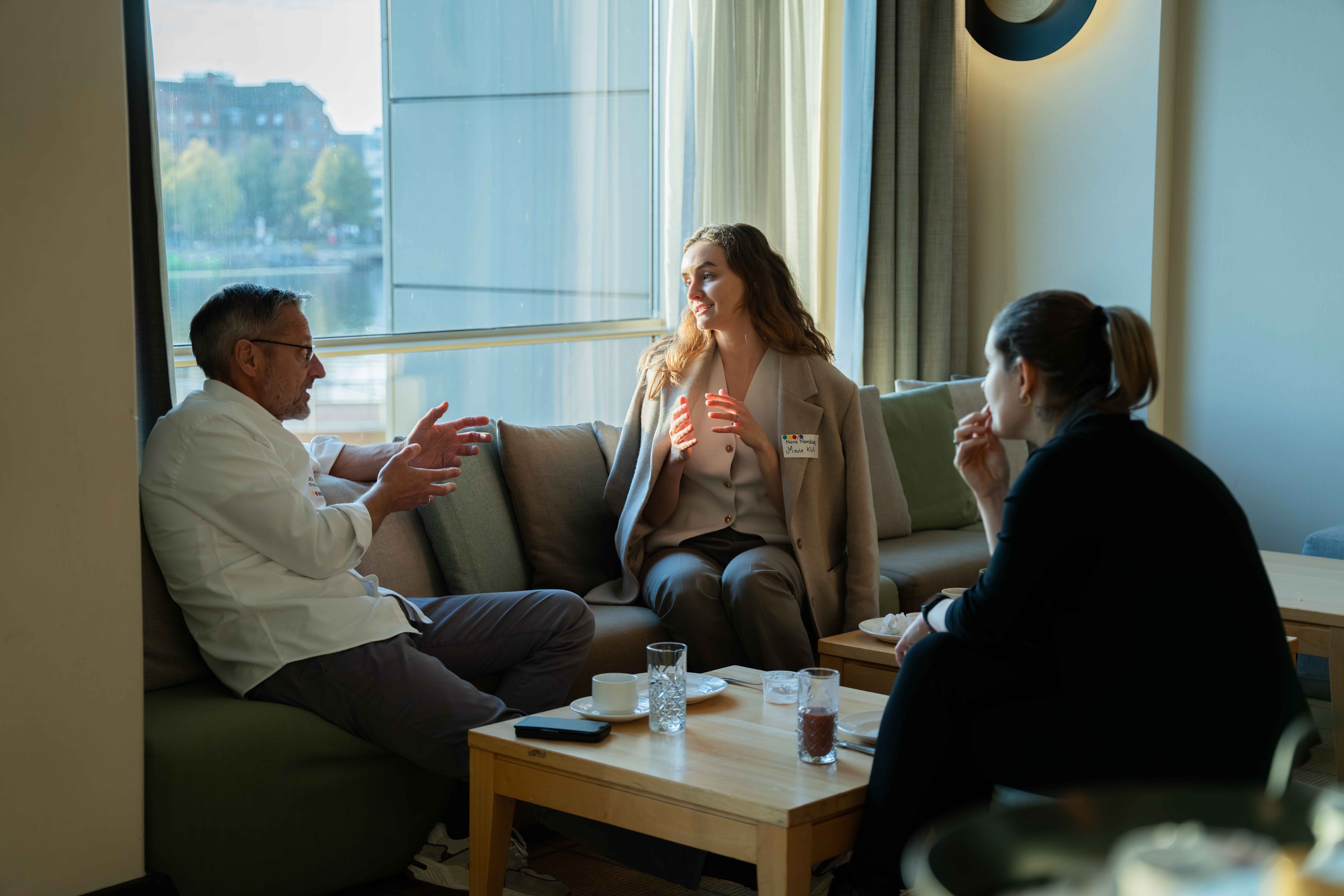 Three people having a conversation while sitting on a couch and chairs around a wooden coffee table in a room with large windows.
