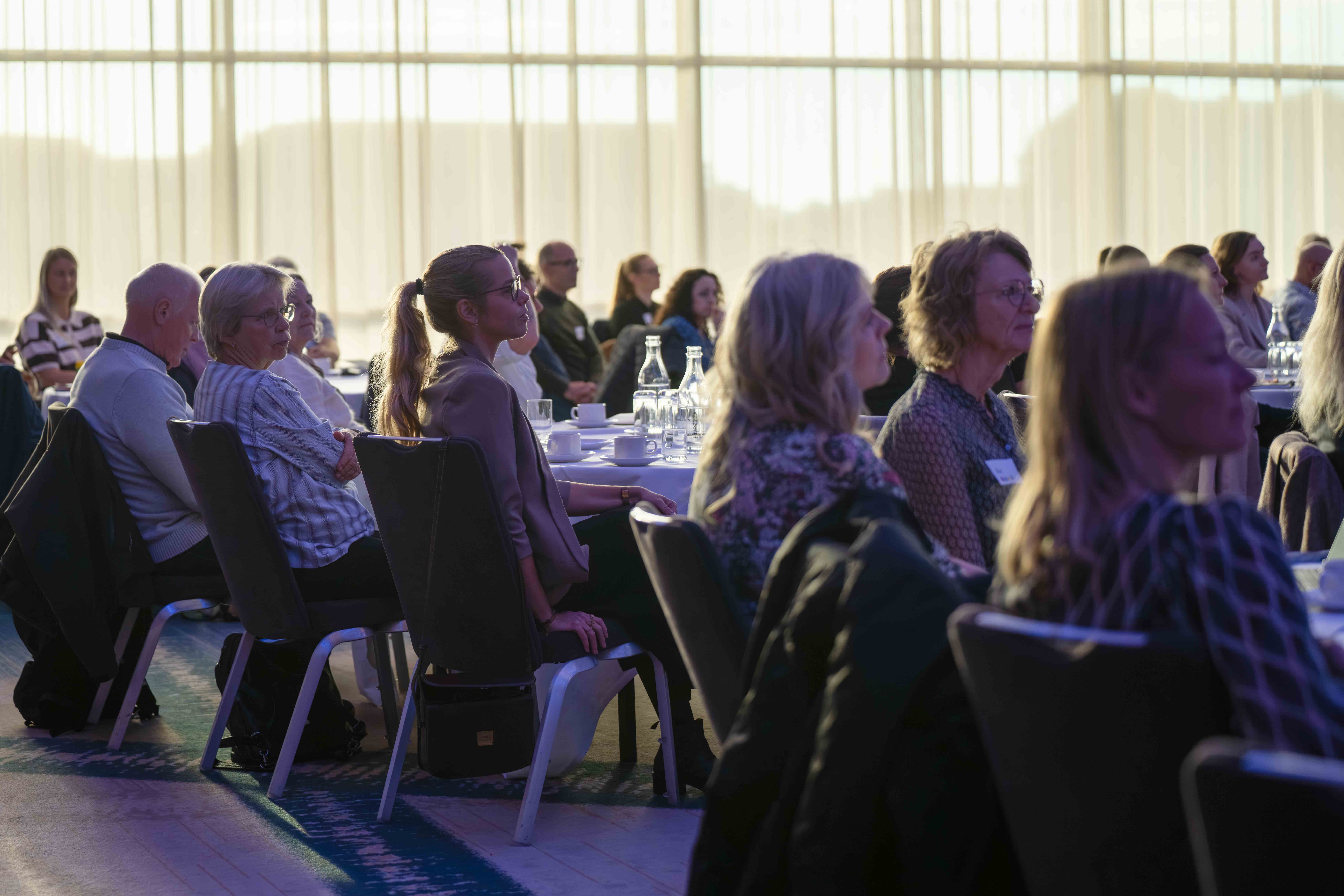 Adults seated in chairs attentively listening at an indoor conference or seminar with tables and glass bottles.