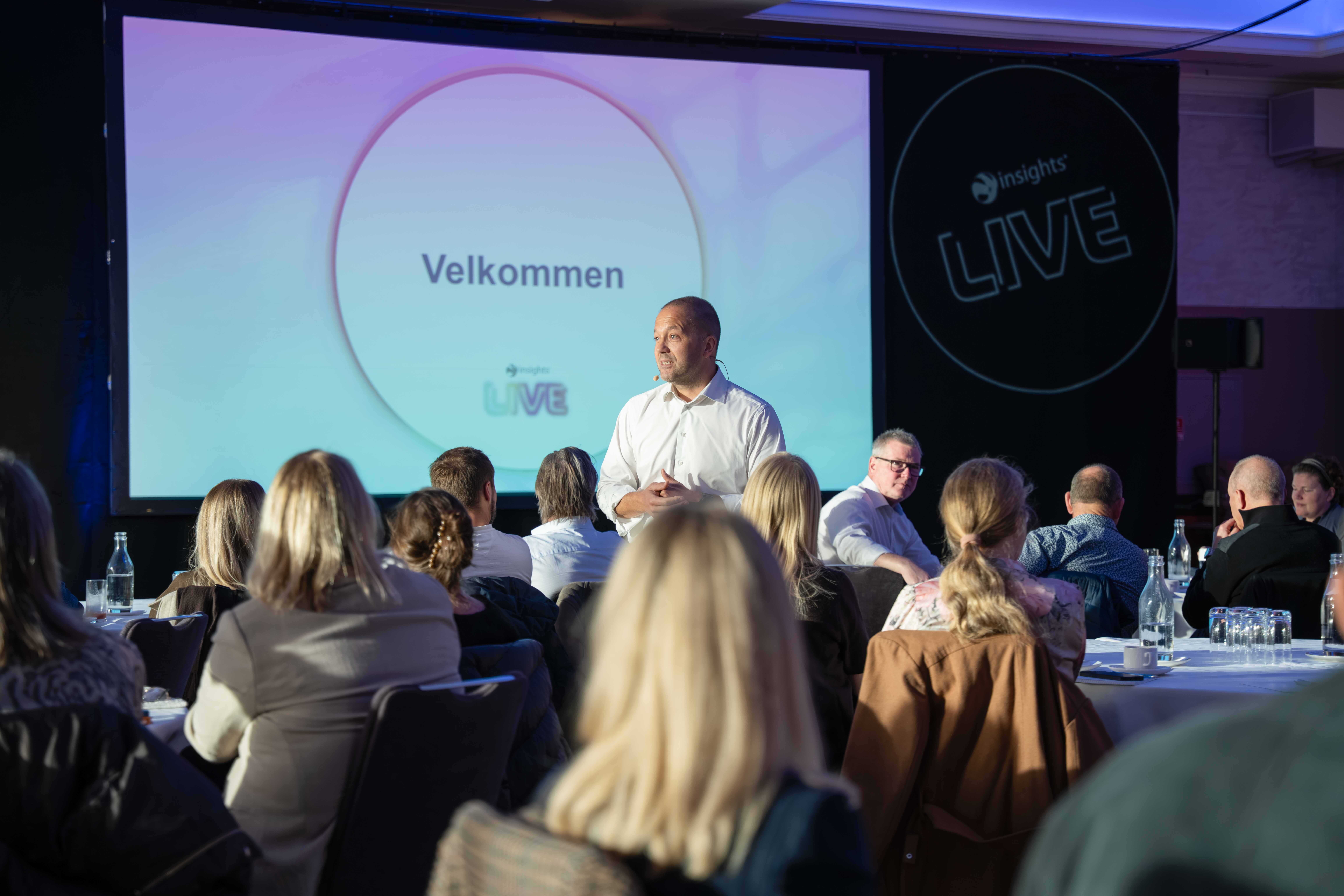 Man in white shirt speaking to an audience at a conference with a presentation screen displaying 'Velkommen' behind him.