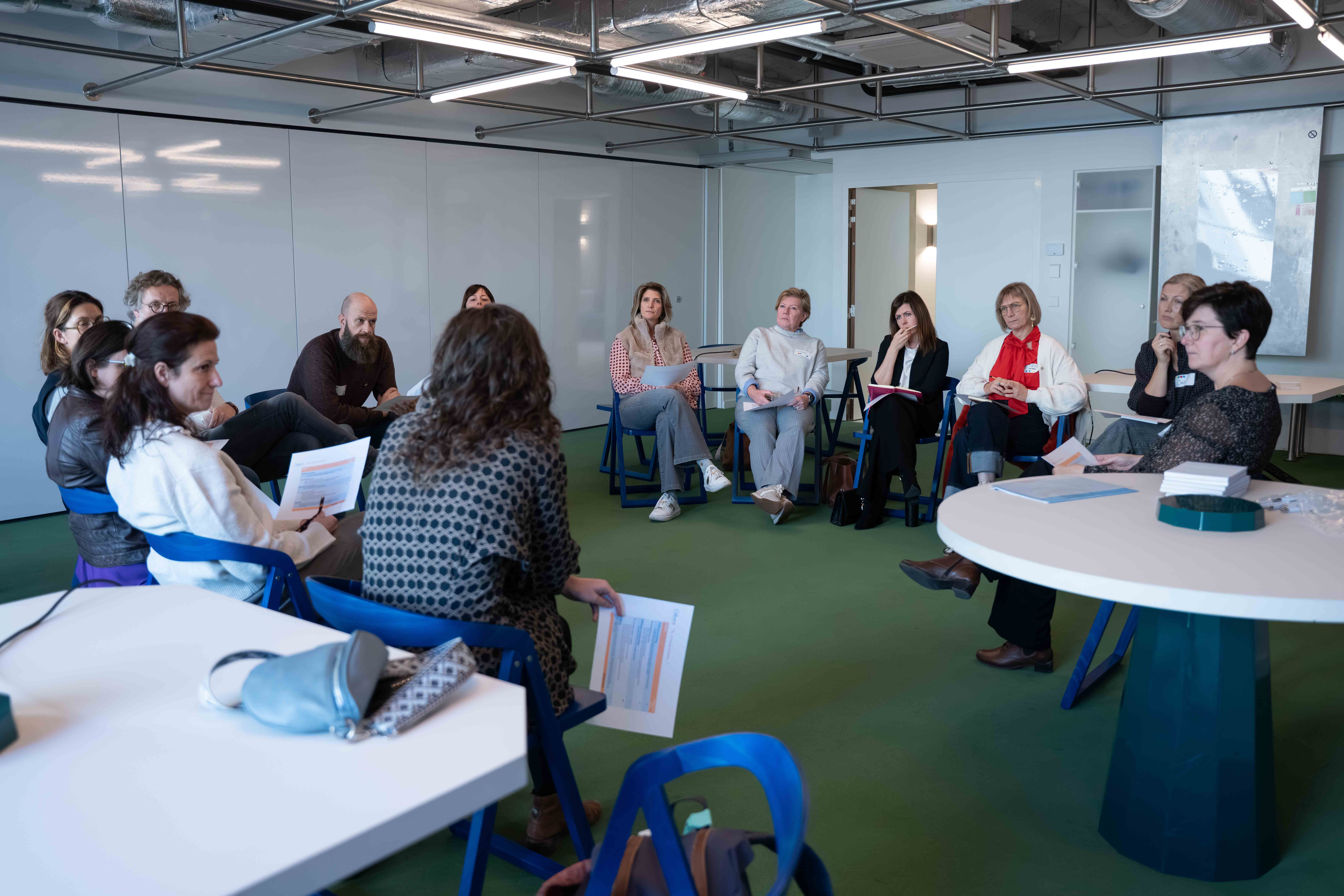 A group of people sitting in a circle in a meeting room, engaged in discussion, holding papers and listening attentively.