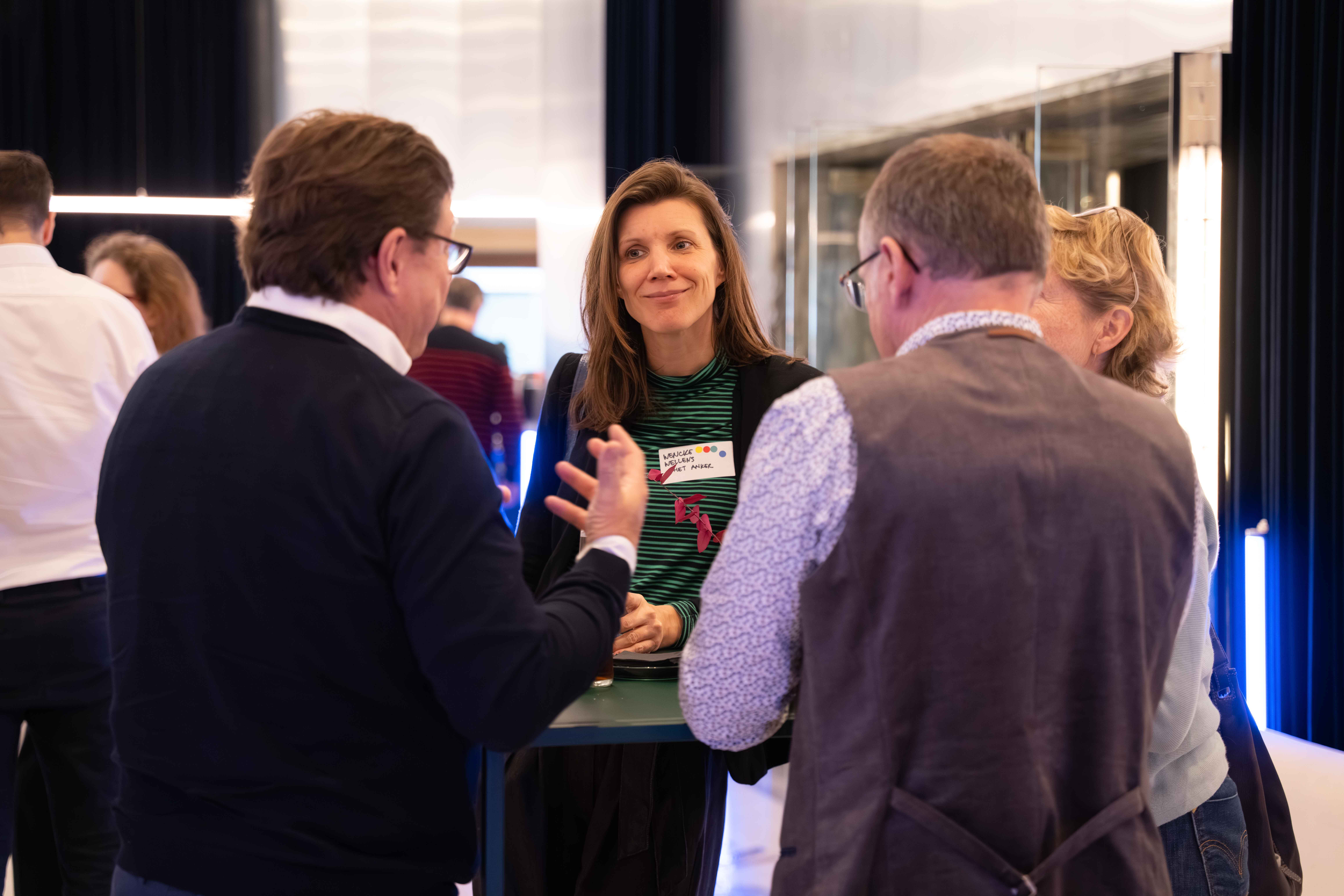 A woman wearing a name tag listens attentively while three people stand around a high table engaged in conversation at an indoor event.