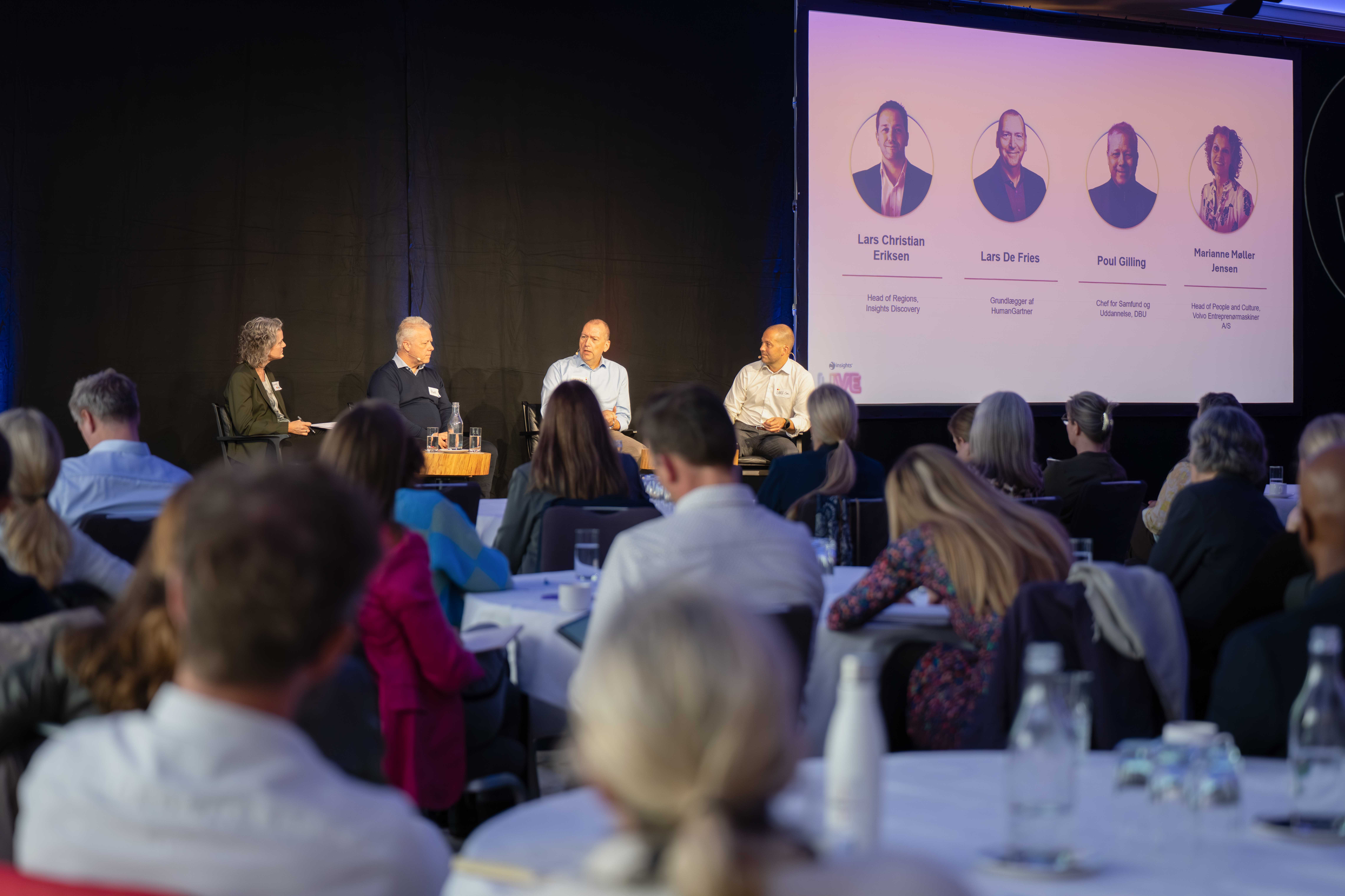 Panel discussion with four speakers on stage and audience seated at round tables.