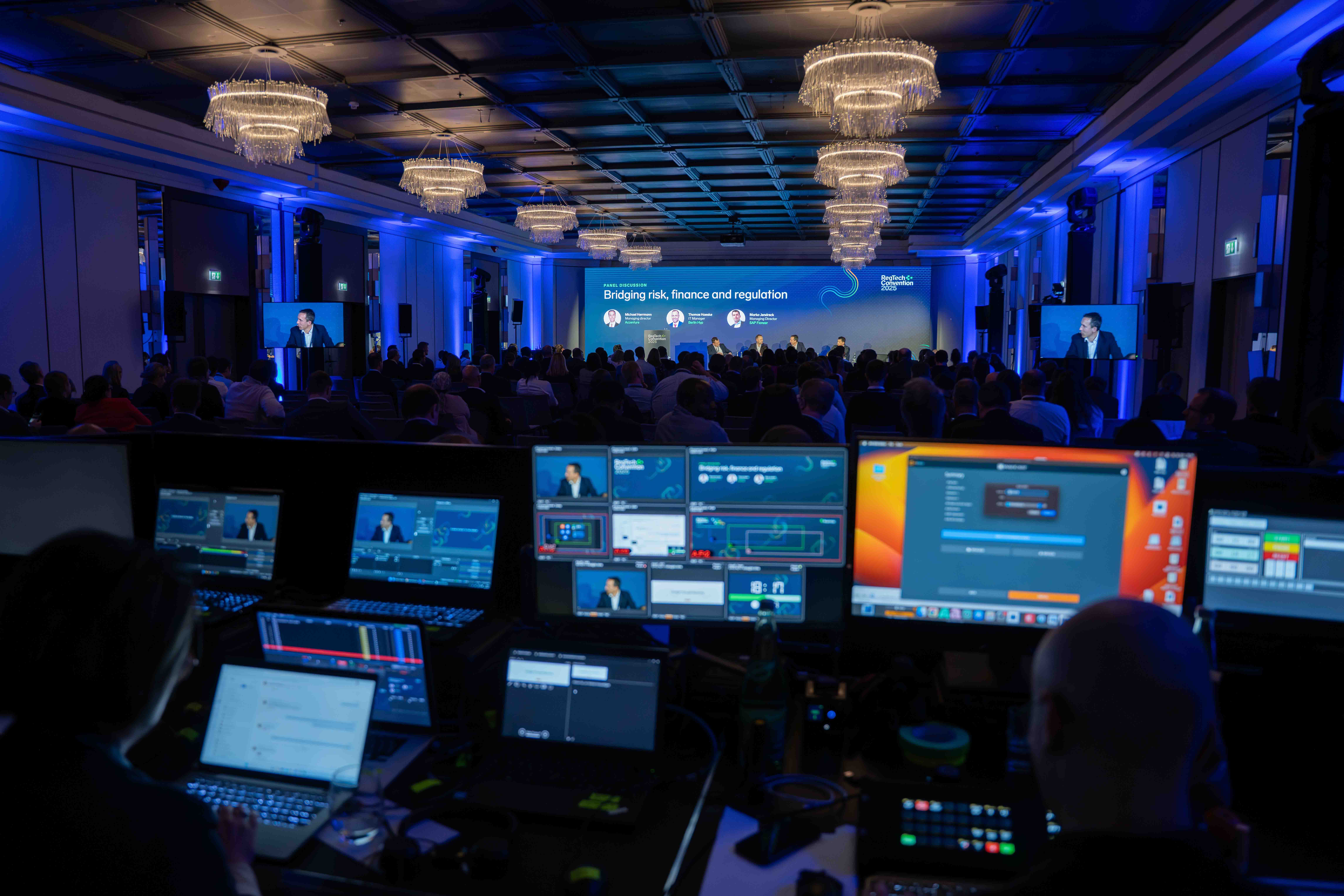 Control room with multiple screens at a conference showing a panel discussion titled 'Bridging risk, finance and regulation' with attendees seated in a large room with chandeliers.