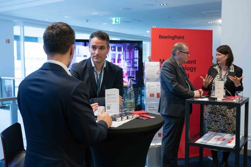 Two pairs of professionals in business attire engaged in discussion at a conference booth with BearingPoint branding.