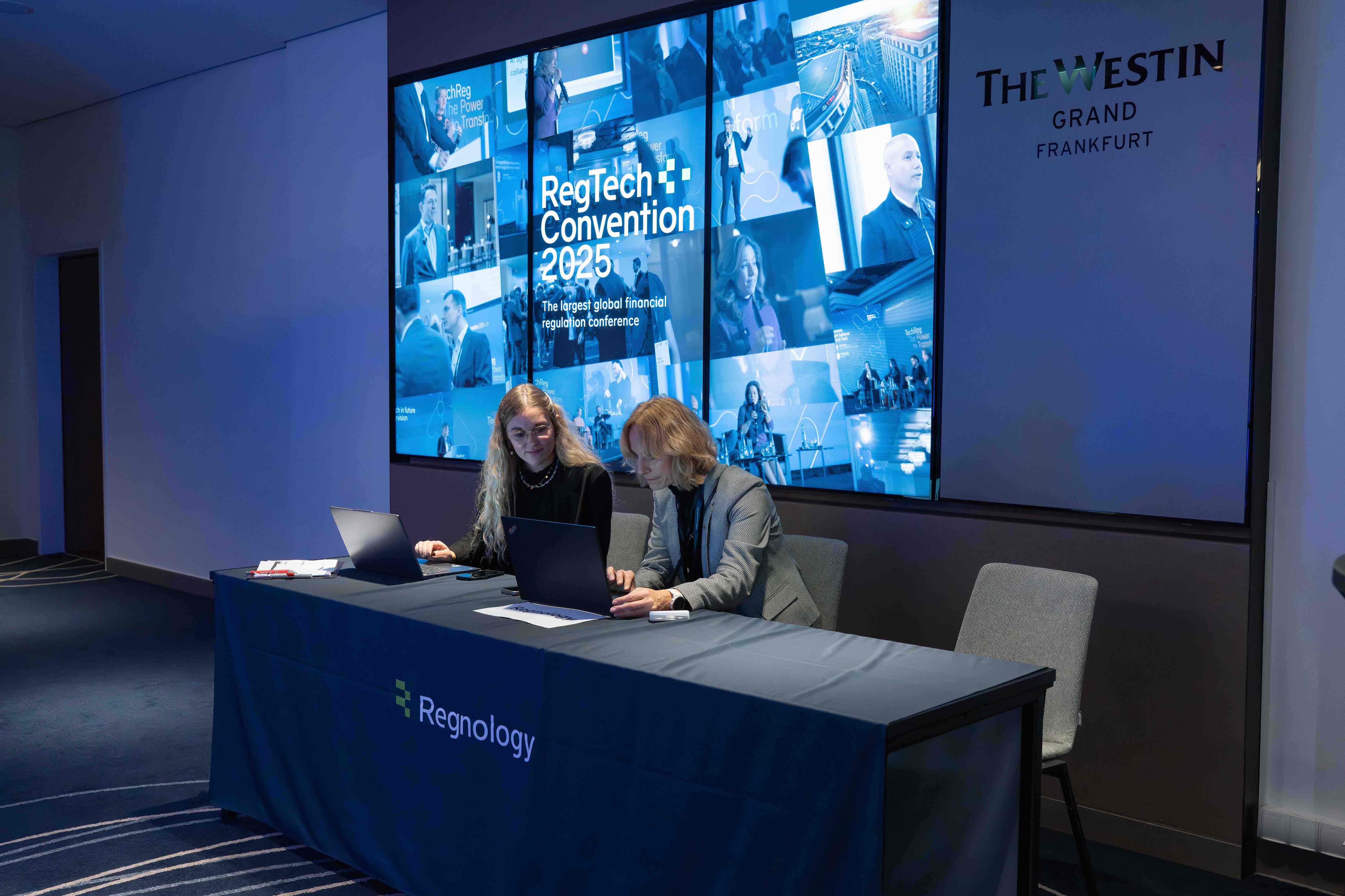 Two women working on laptops at a Regnology table in front of a large screen displaying RegTech Convention 2025 at The Westin Grand Frankfurt.
