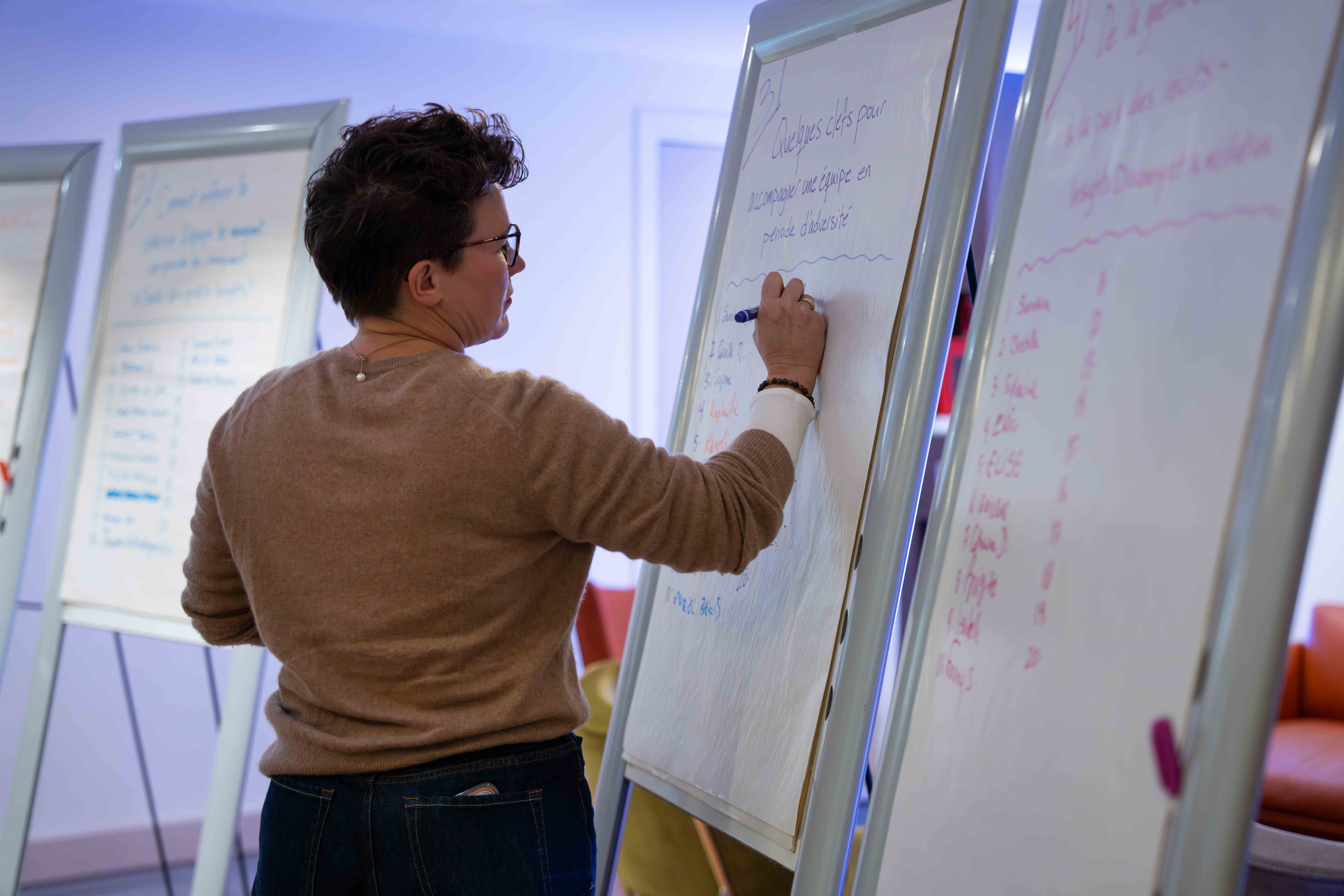 Person with short dark hair and glasses writing notes on a large white flip chart in a conference room.