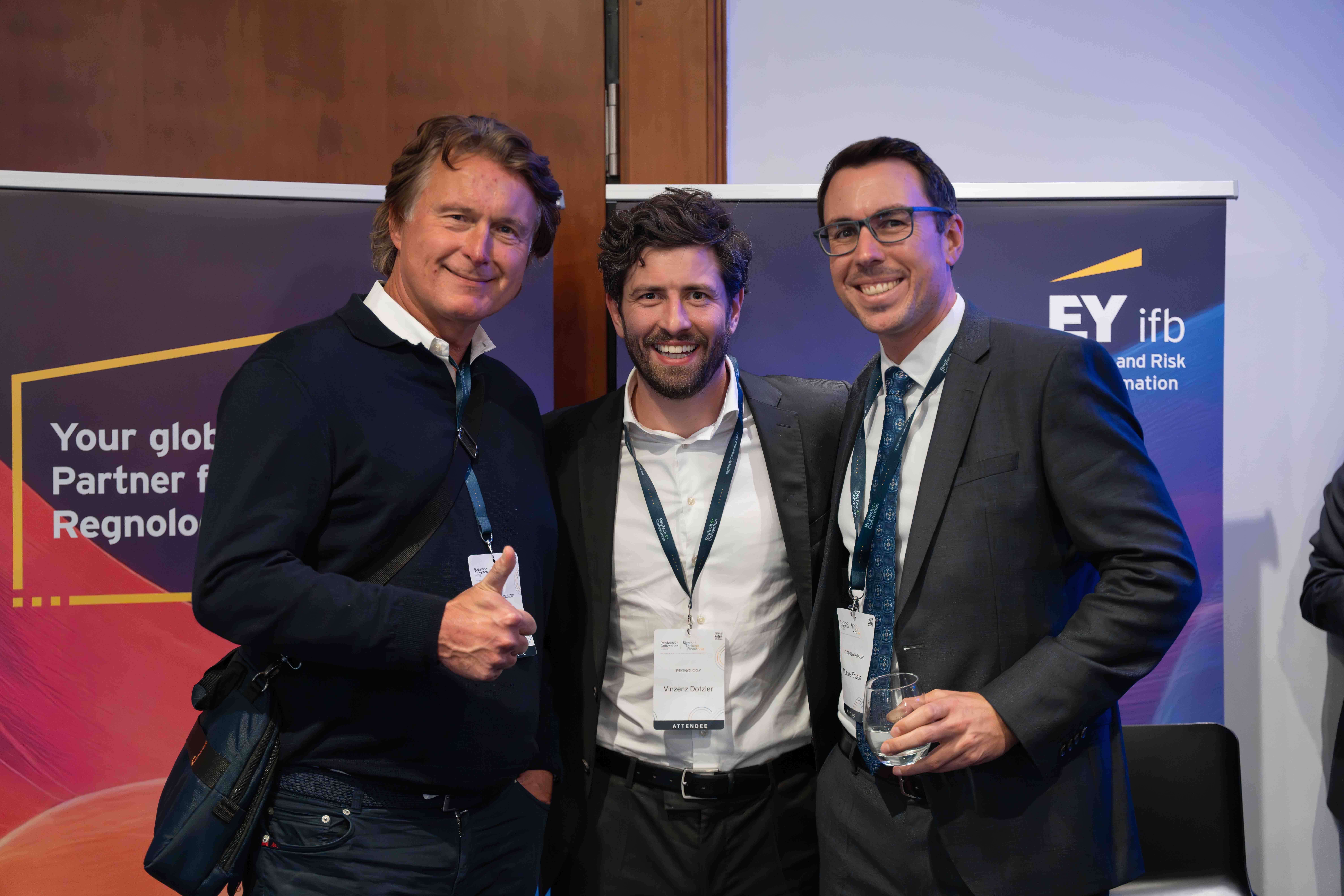 Three men smiling and posing together at a professional event, two wearing suits and one in a dark sweater, with conference badges and an EY branded backdrop.