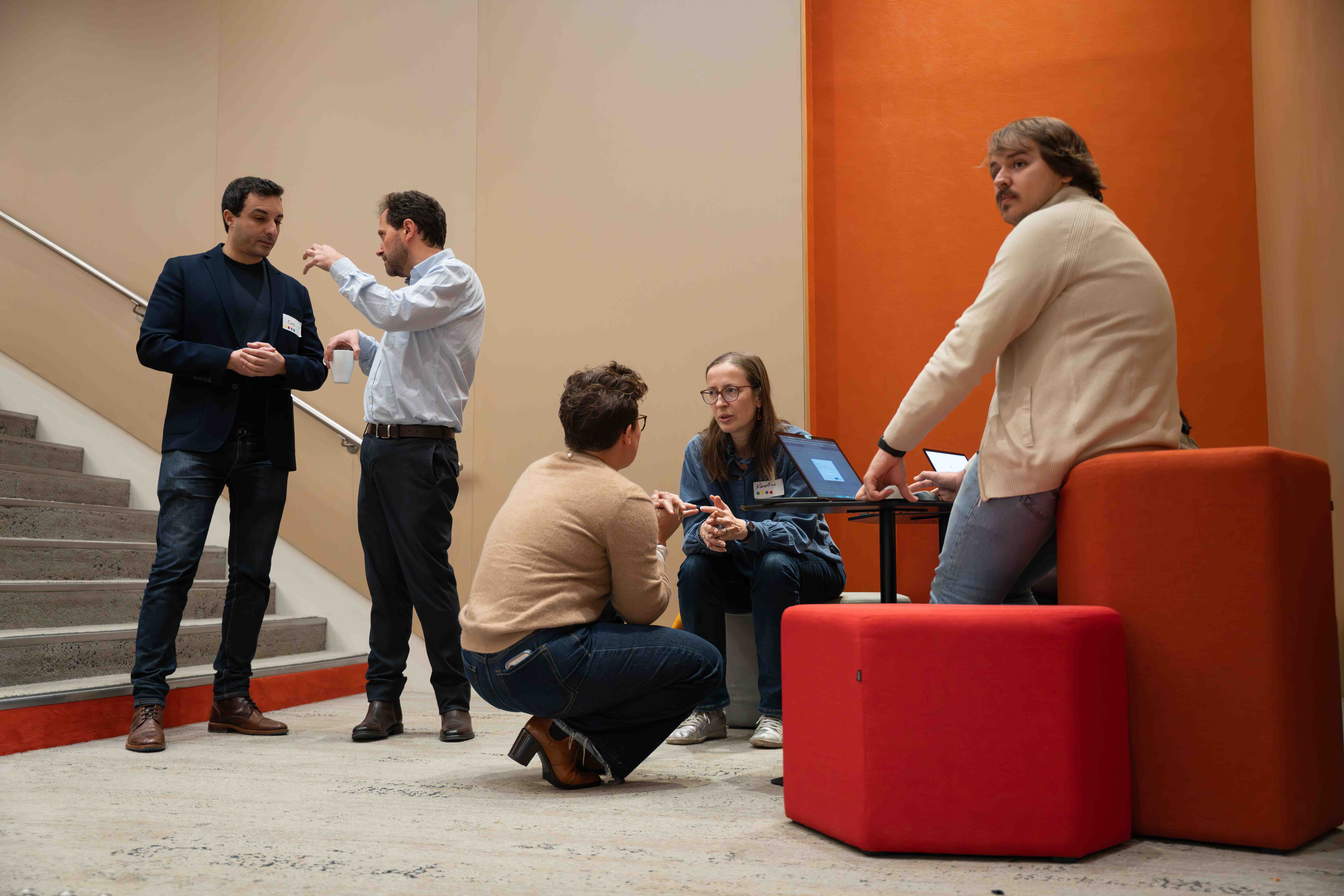 Five people in a casual business setting having conversations; two men standing by stairs, two women seated or crouched near laptops, and one man sitting on a red stool looking back.