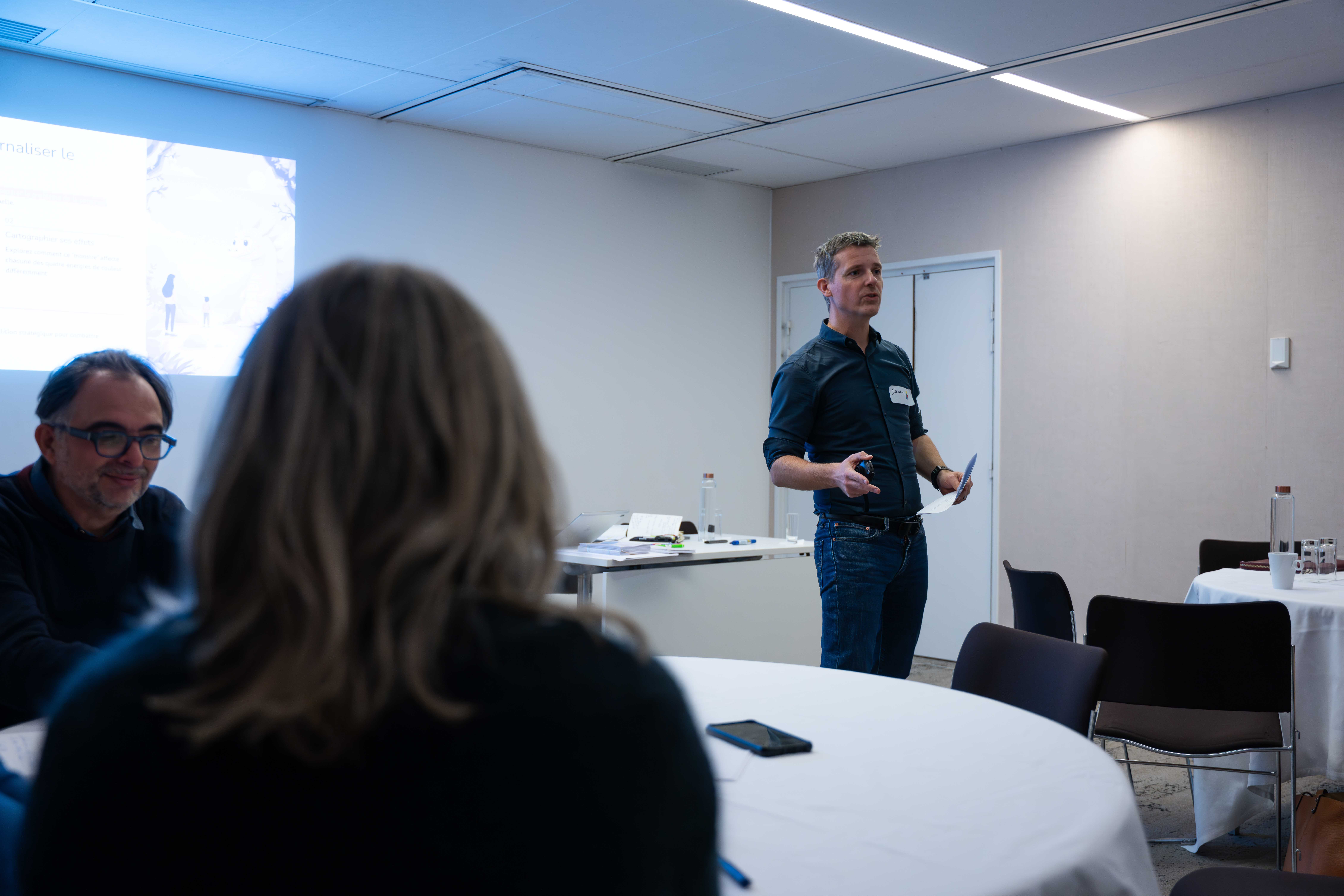 Man standing and speaking in a small conference room with people seated around round tables.