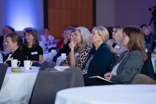 Audience of attentive women seated at round tables in a conference room with white tablecloths and cups.