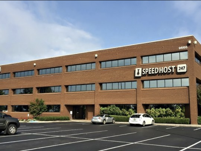 A three-story brick office building with large windows, a "Speedhost 247" sign, and a nearly empty parking lot with a few cars parked in front. The sky is partly cloudy.