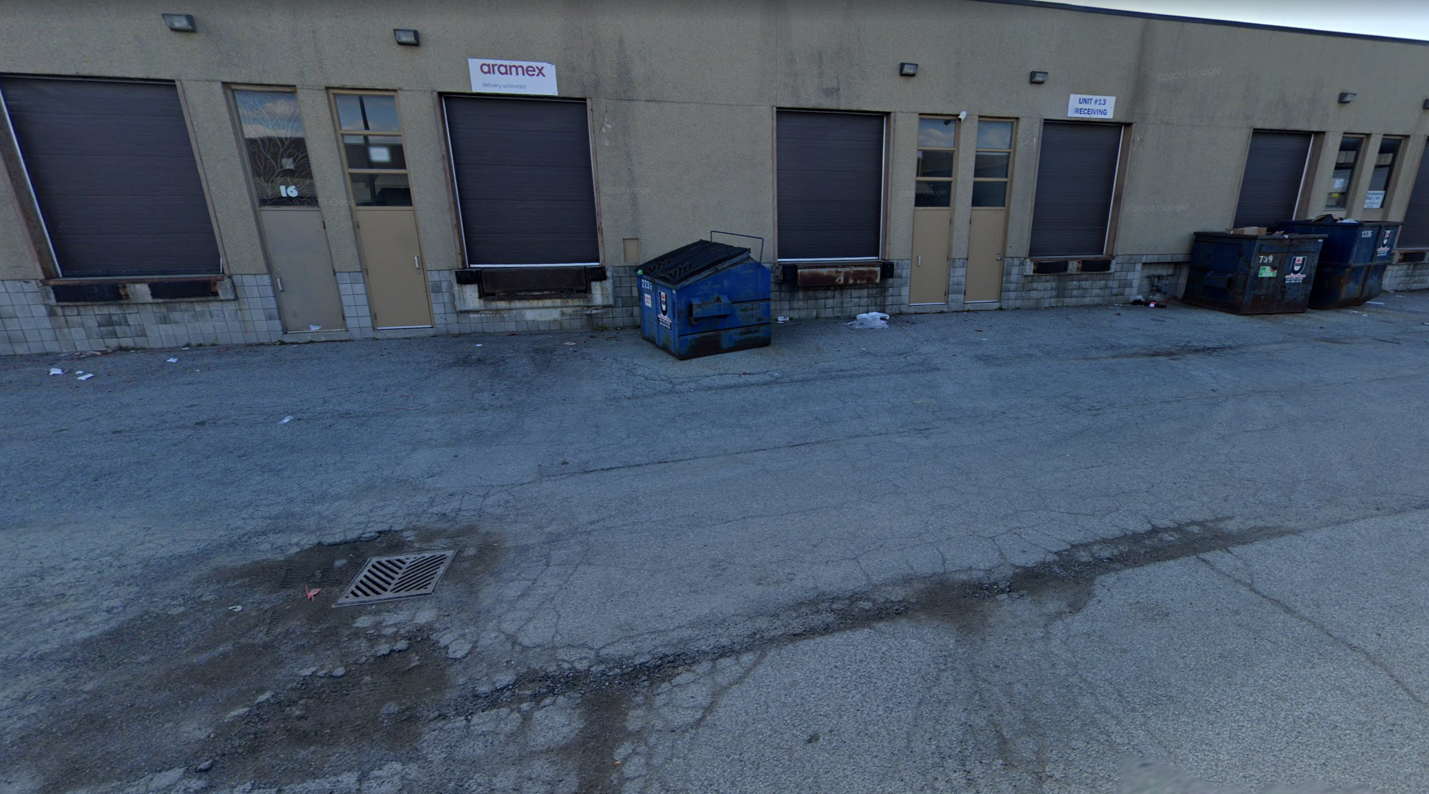 A loading dock with several closed garage doors, labeled "aramex" and "WTC Logistics," and two blue dumpsters. The ground is cracked and littered with some trash. The area appears industrial and deserted.