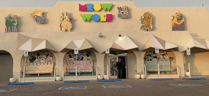 Front entrance of Meow Wolf art installation with colorful sign, murals, and unique sculptures above the building.