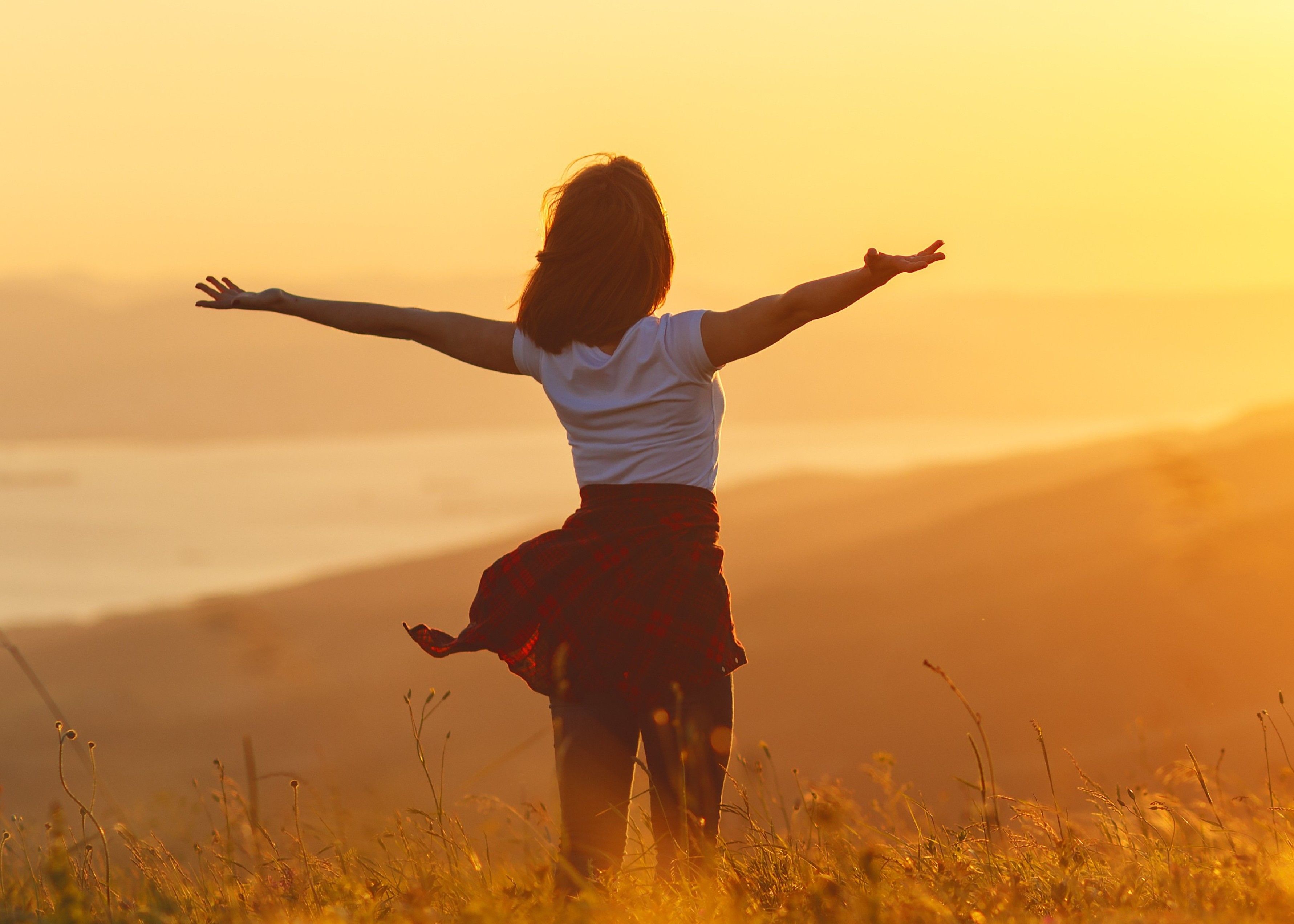 Person standing in a field with arms outstretched at sunset, overlooking distant hills and water.