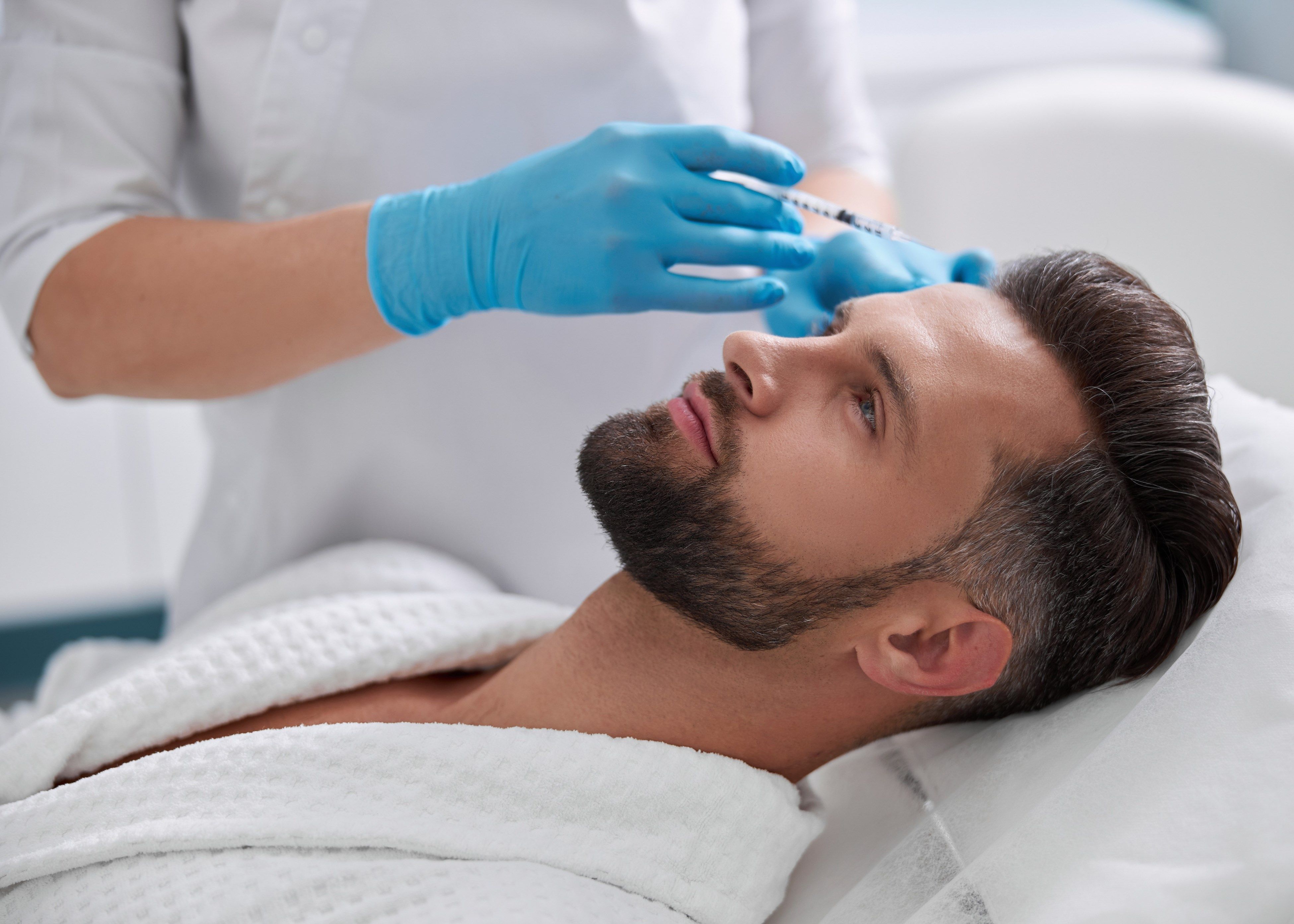 Man in a white robe lying down receiving a cosmetic injection in the forehead from a professional wearing blue gloves.