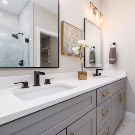 Modern bathroom with a double sink vanity featuring gray cabinets, black faucets, gold drawer handles, two mirrors, and a glass-enclosed shower.