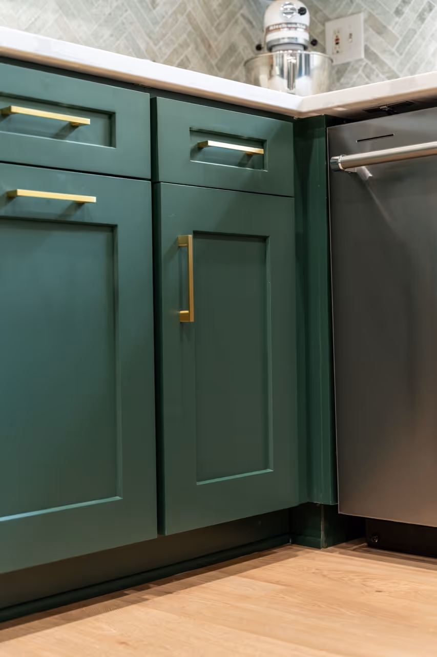 Close-up of green kitchen cabinets with brass handles next to a stainless steel dishwasher under a white countertop.