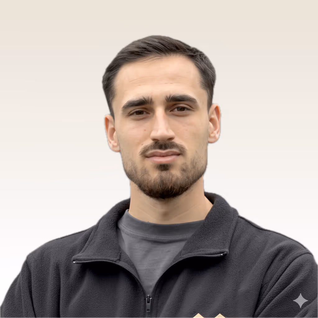 Portrait of a young man with short dark hair and a beard wearing a black fleece jacket over a gray shirt against a light gradient background.