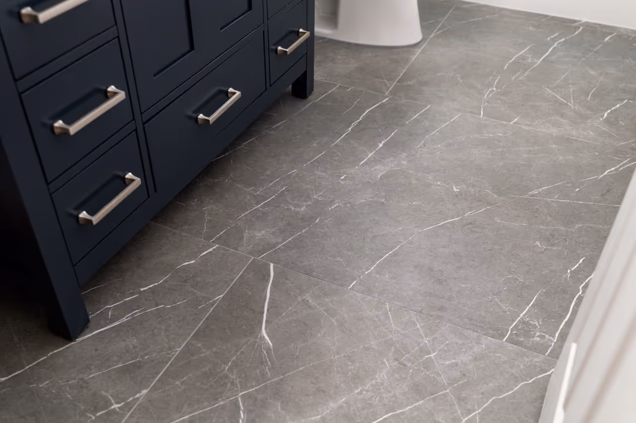 Close-up of gray marble floor tiles with white veining next to dark blue cabinet drawers and a white toilet base in a bathroom.