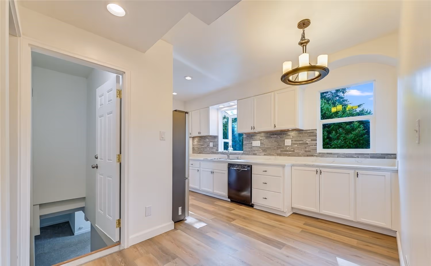 Bright kitchen with white cabinets, a gray tiled backsplash, hardwood floors, black dishwasher, and a window showing green trees.