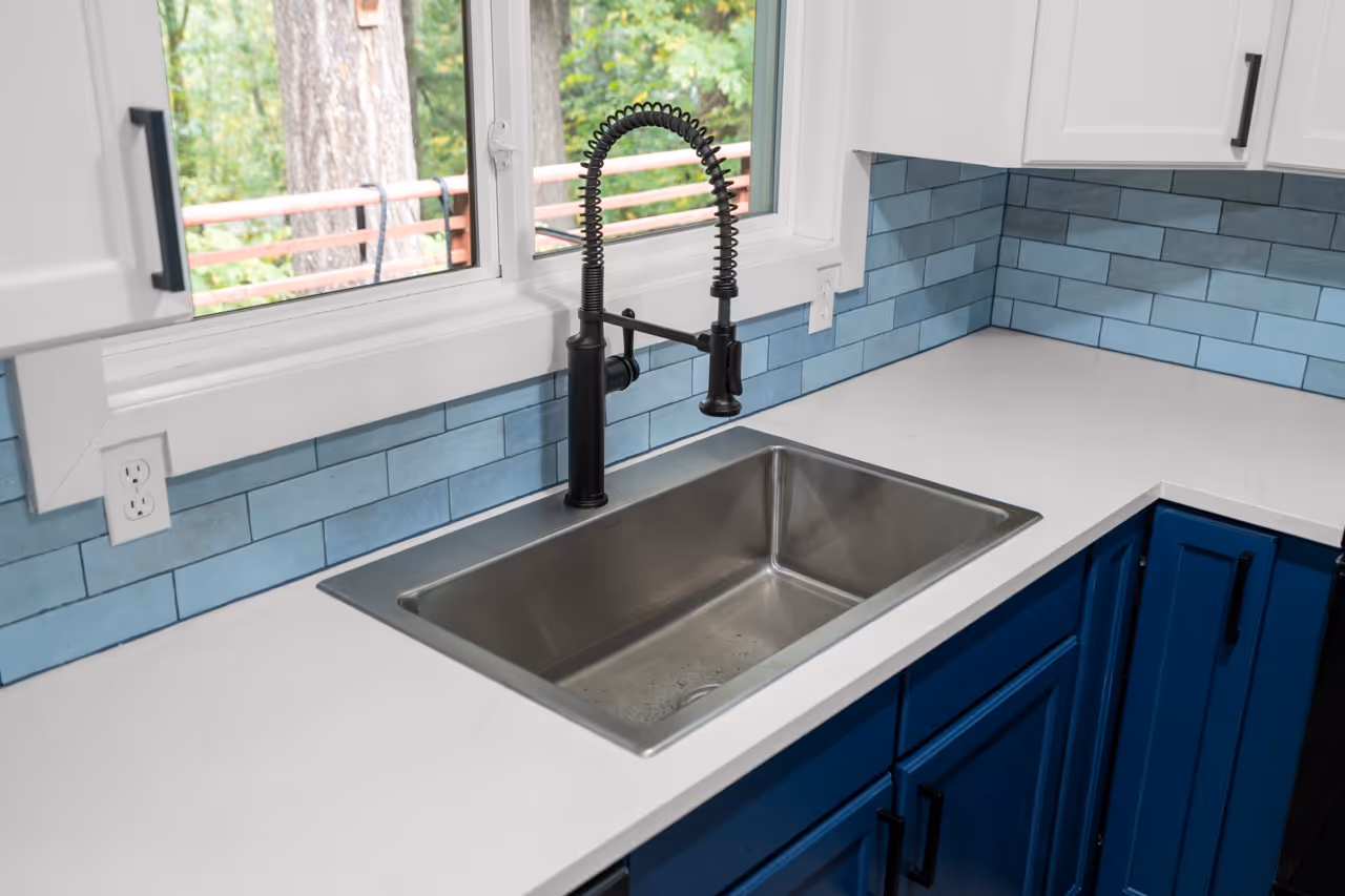 Modern kitchen sink with a black spring coil faucet set into a white countertop with blue subway tile backsplash and blue cabinets.