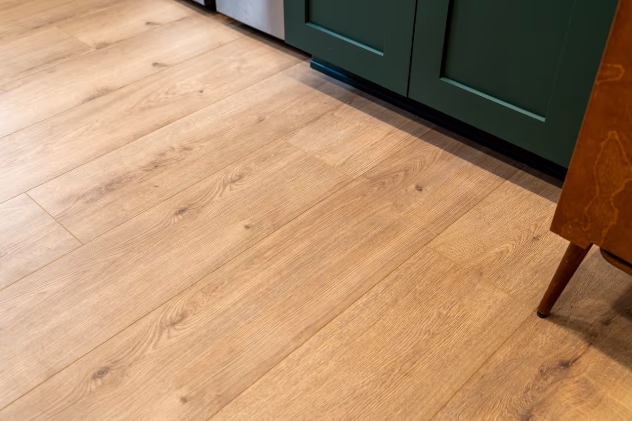 Light wood plank flooring with natural grain next to green lower kitchen cabinets and a mid-century wooden furniture leg.