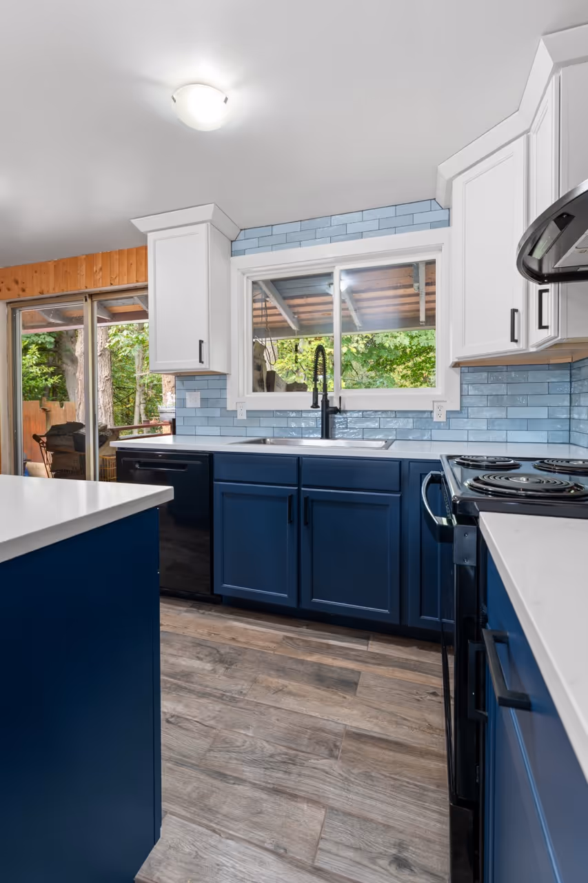 Modern kitchen with navy blue lower cabinets, white upper cabinets, light blue subway tile backsplash, wood-look floor, and a large window above the sink.