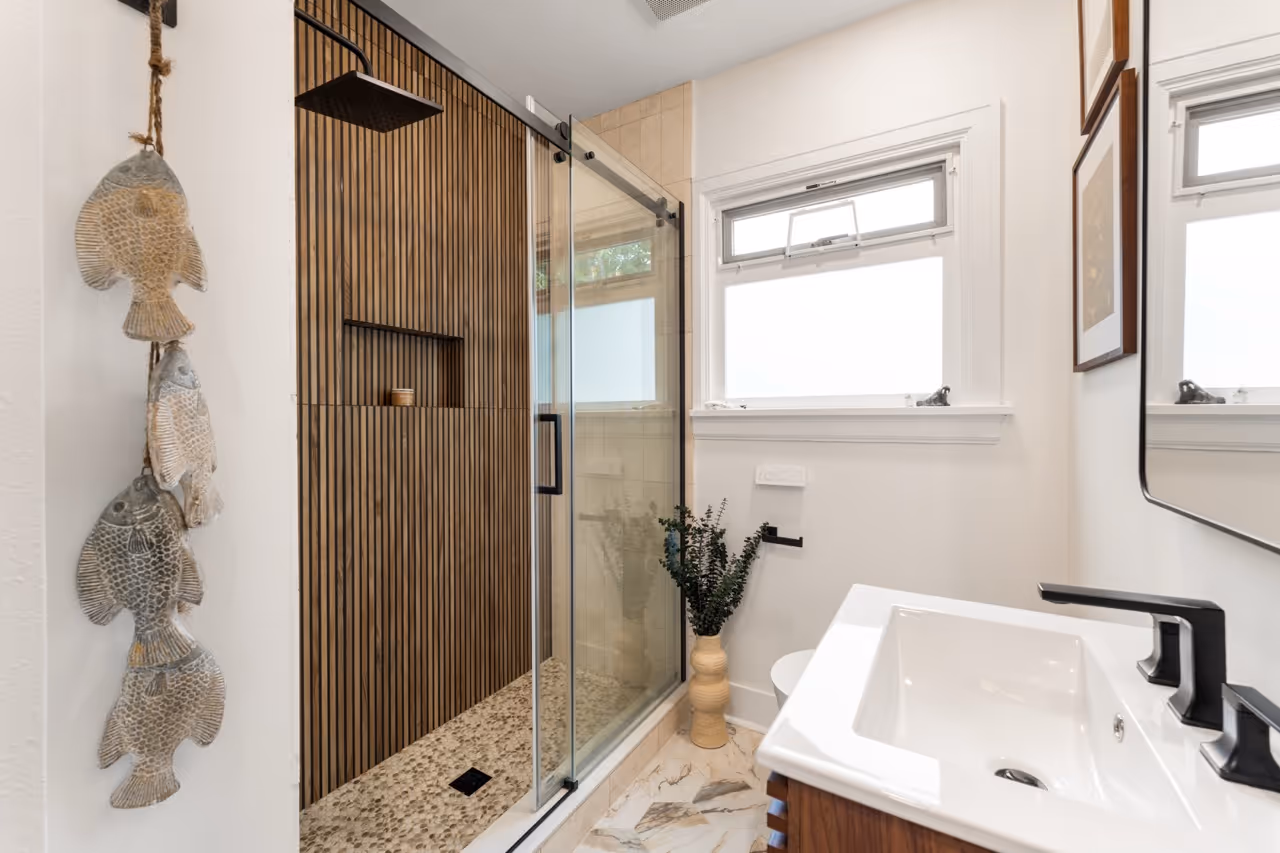 Modern bathroom with a glass shower door, pebble floor, vertical wood paneling, white sink with black faucet, and decorative fish hanging on the wall.