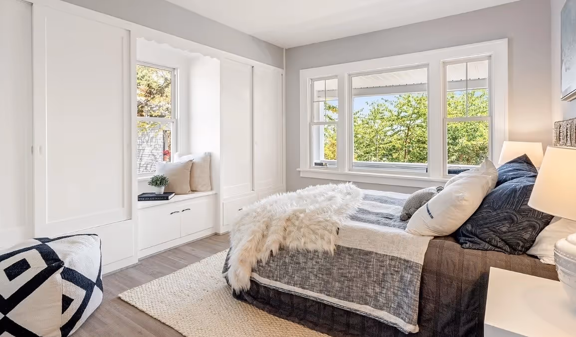 Bright bedroom with large windows, a bed with dark gray and white bedding, a fluffy throw, and a built-in window seat with cushions and a small plant.
