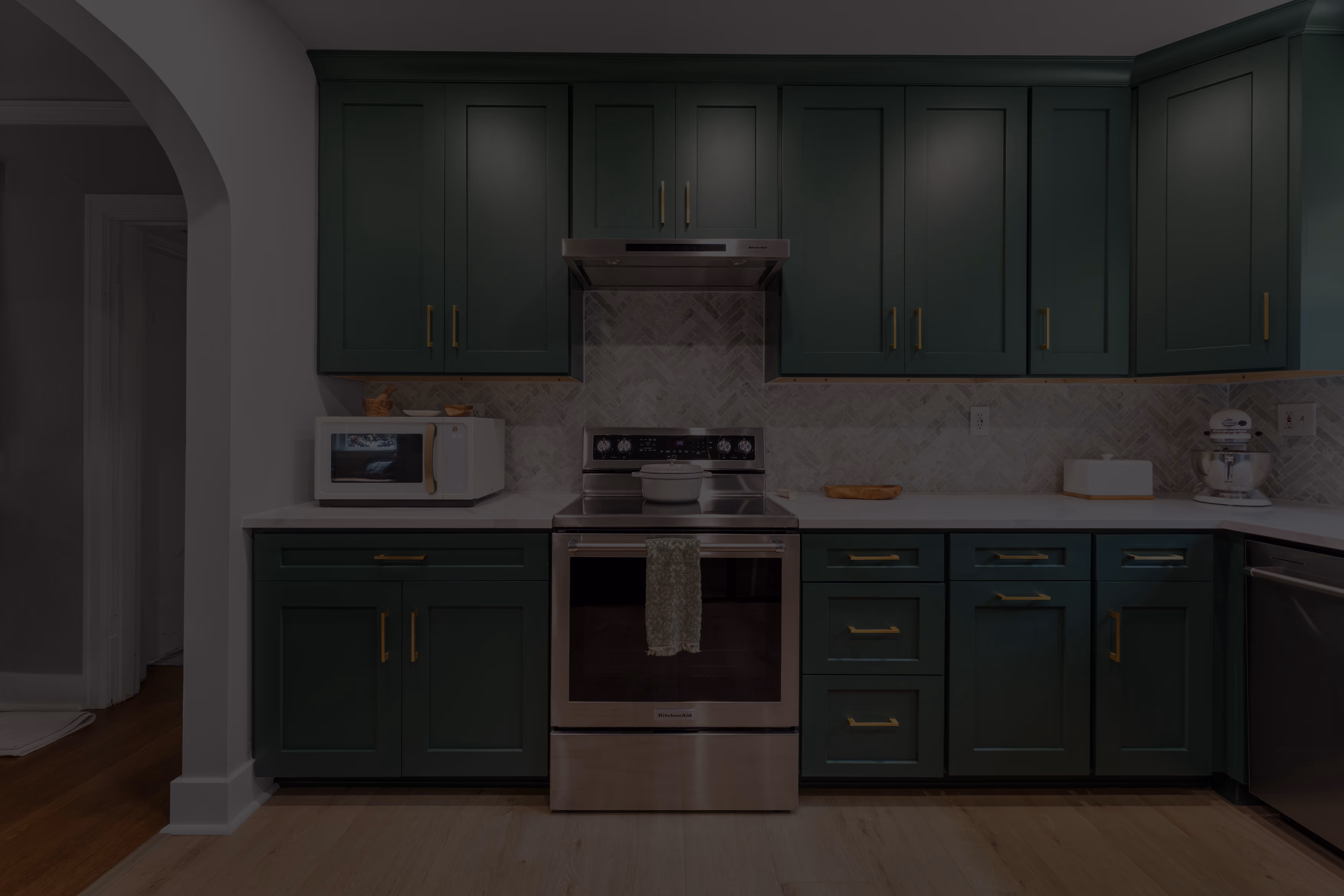 Modern kitchen with dark green cabinets, stainless steel stove with pot, microwave on the left, and a stand mixer on the right.