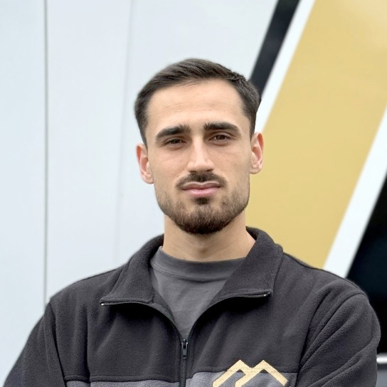 Young man with short dark hair and beard wearing a black zip-up jacket, standing against a modern geometric background.