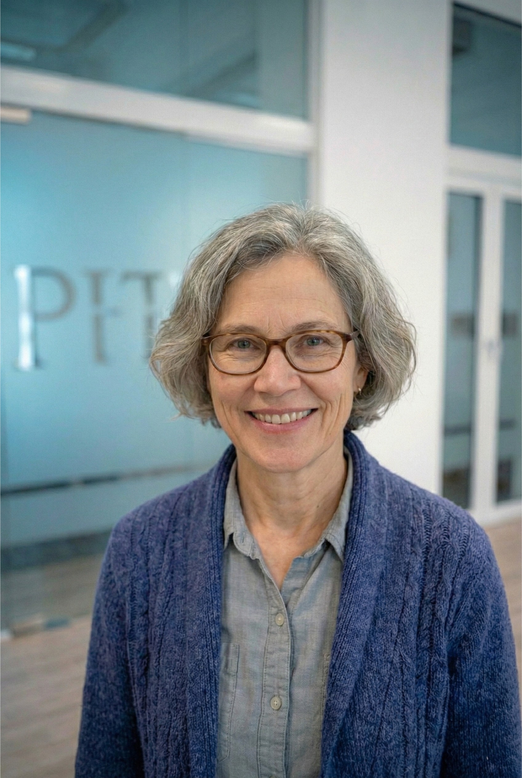 Smiling middle-aged woman with short gray hair and glasses wearing a blue cardigan and gray shirt in an indoor office setting.