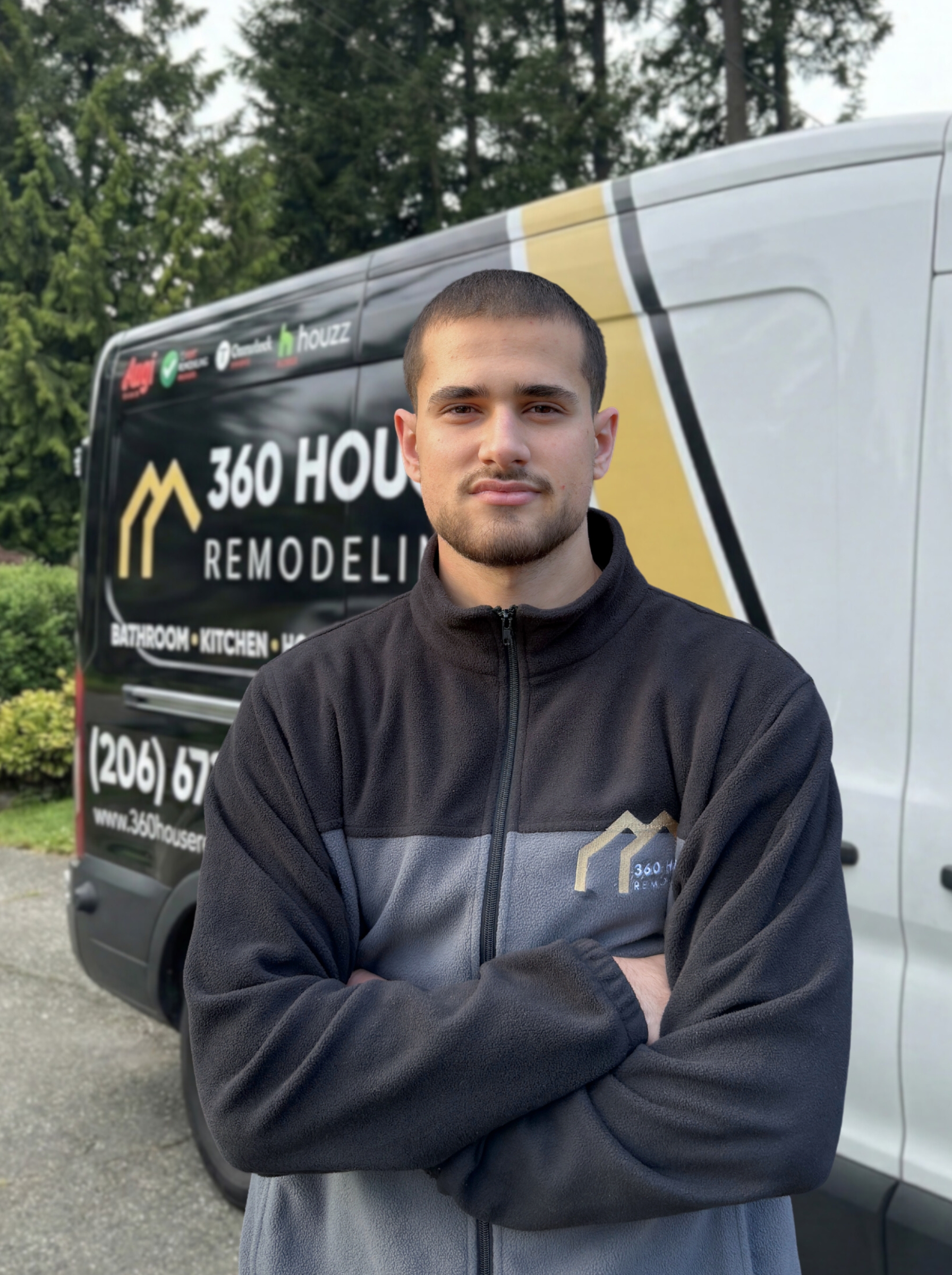 Young man with short hair and beard wearing a black and gray zip-up fleece, standing with arms crossed in front of a 360 House Remodeling van.