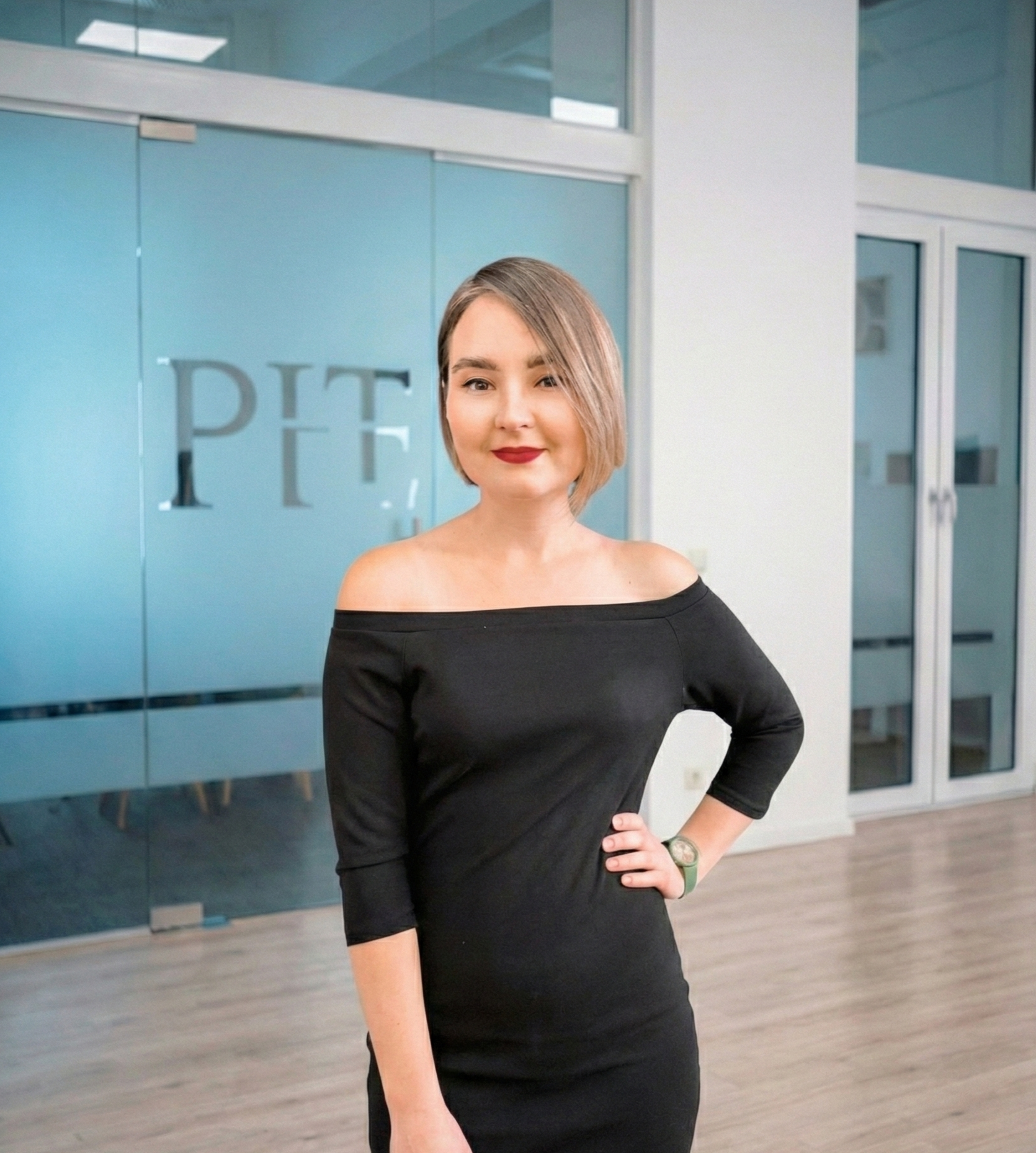 Woman with short blonde hair wearing a black off-shoulder dress, standing with hand on hip in a modern office space.