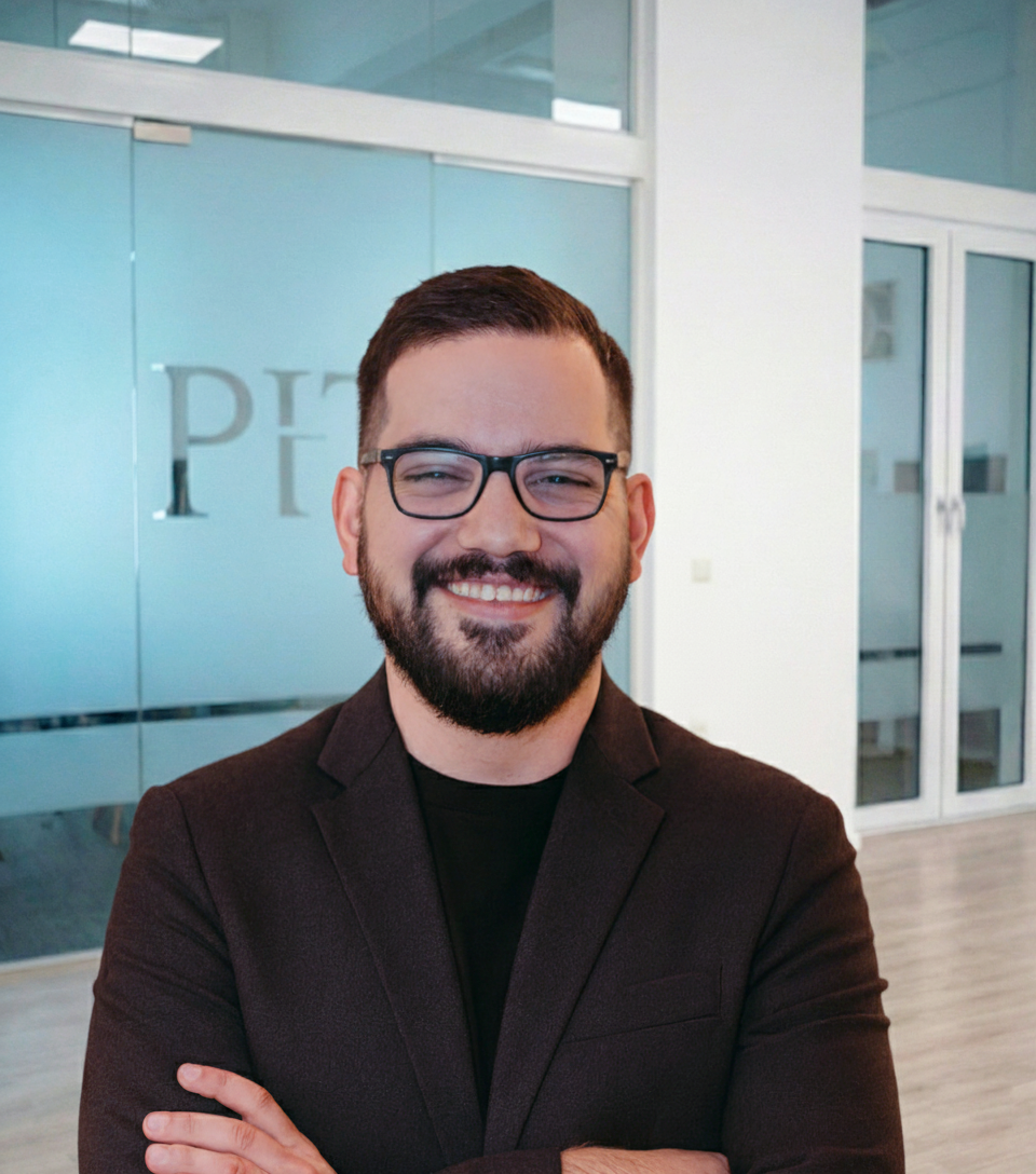 Smiling man with glasses and beard wearing a dark blazer, standing with arms crossed in an office.