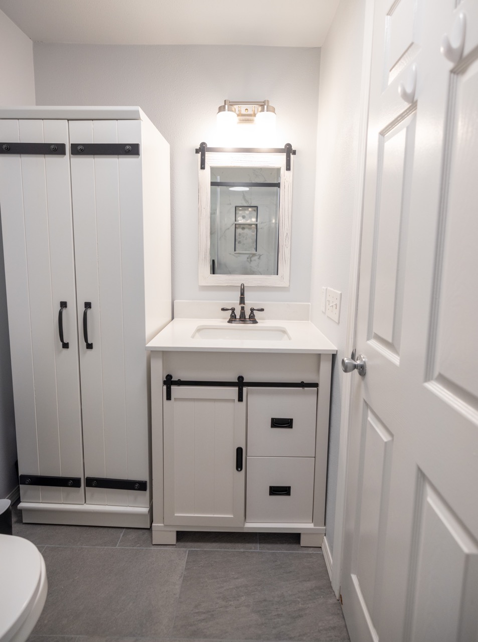 Modern white bathroom vanity with black hardware, rectangular mirror, and light fixture above on gray tiled floor.