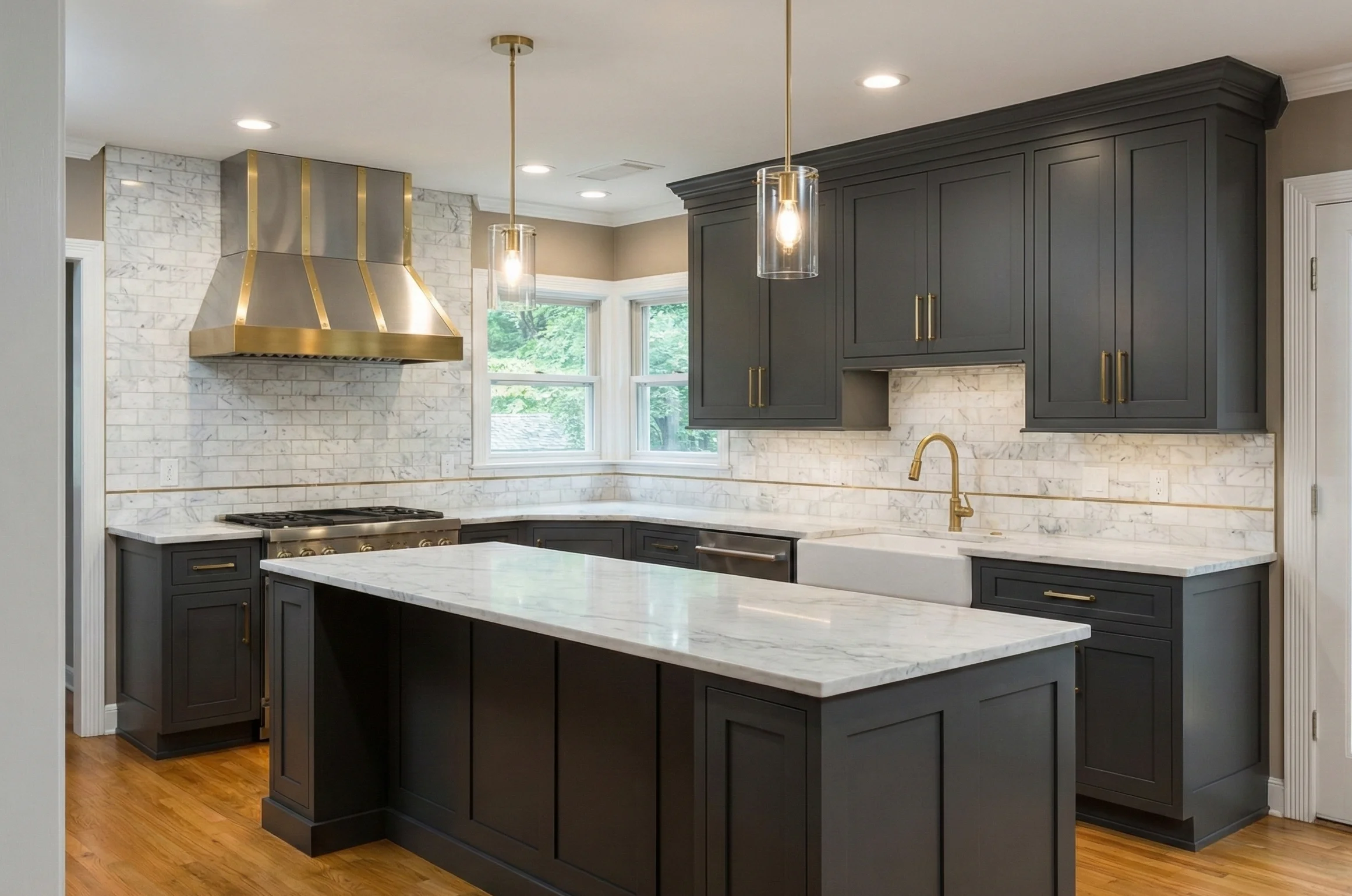 Modern kitchen with dark gray cabinets, marble countertops, brass faucet and range hood, hardwood floor, and two pendant lights.