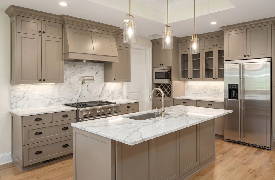Modern kitchen with beige cabinetry, marble countertops and backsplash, stainless steel appliances, and a central island with a gold faucet.