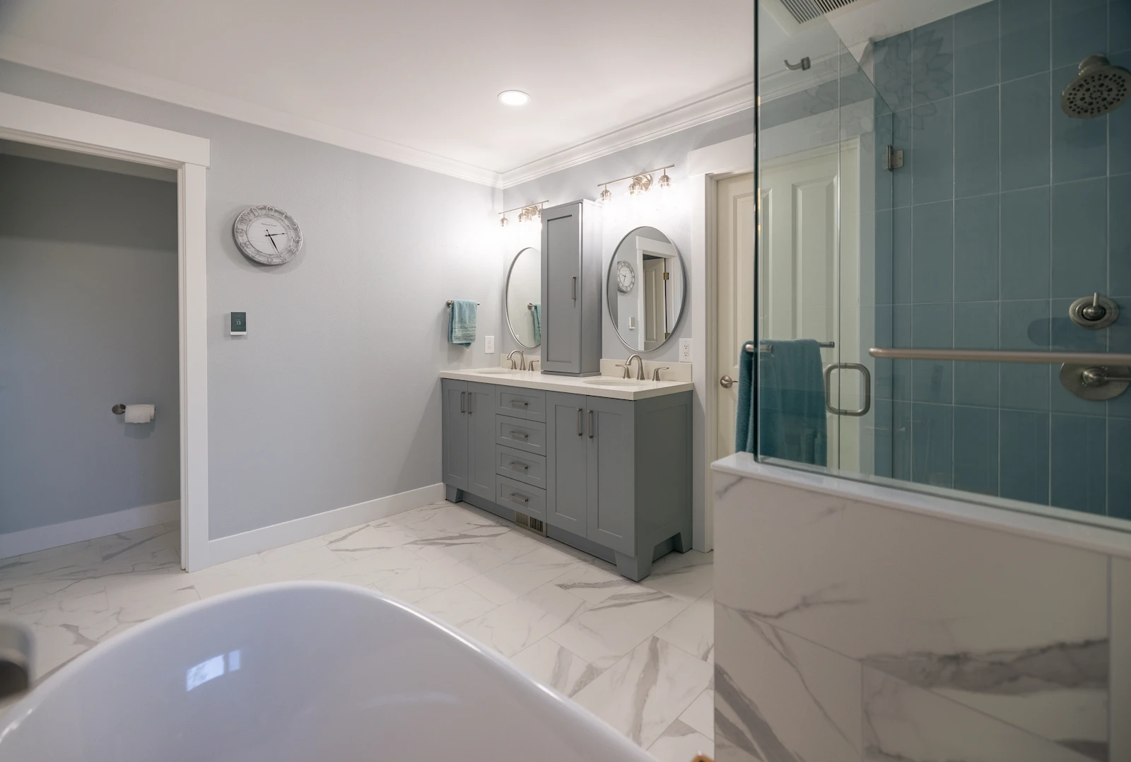 Modern bathroom with gray double-sink vanity, oval mirrors, marble floor tiles, glass shower with blue tiles, and a white freestanding bathtub.