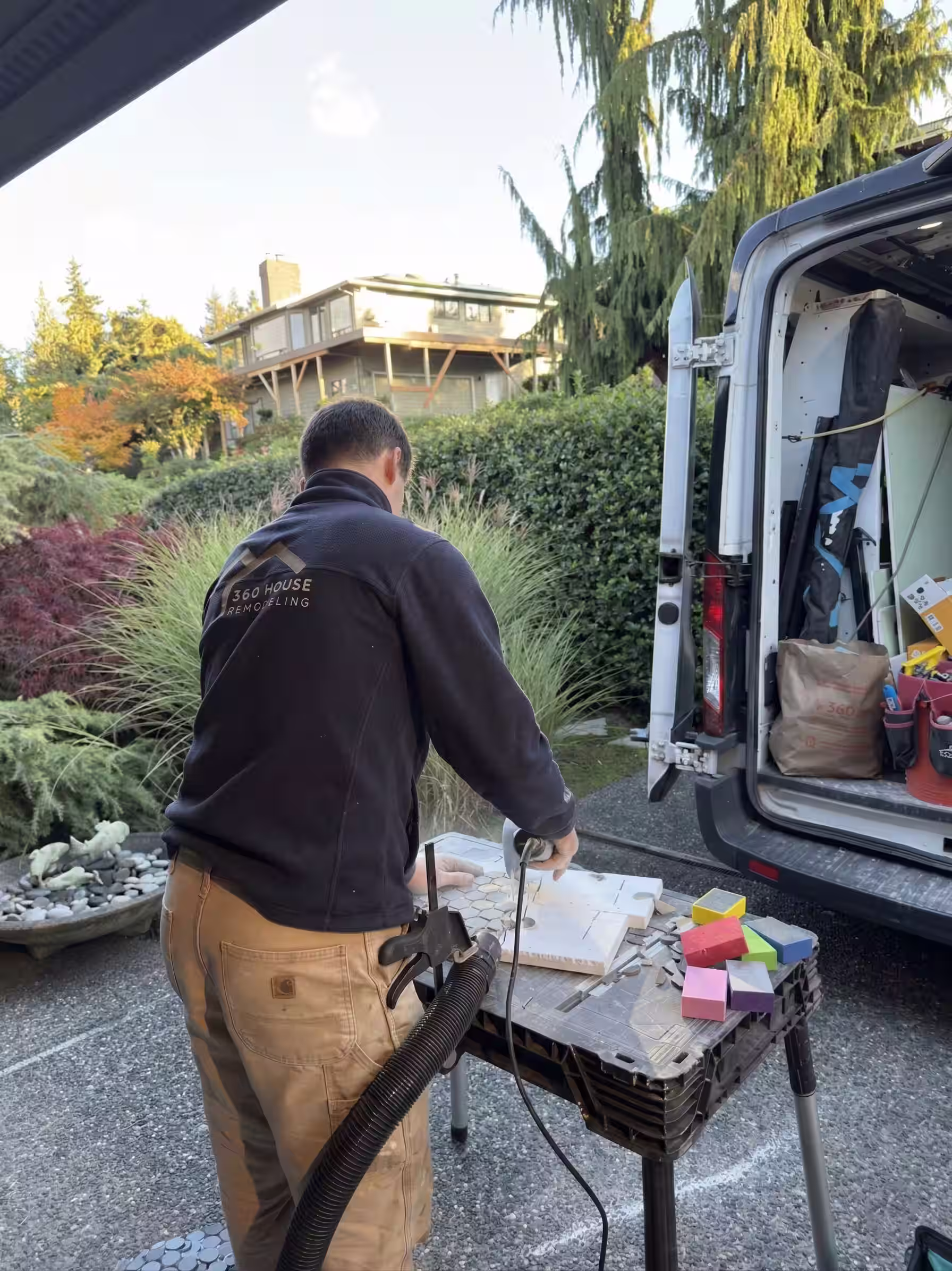 Man working on a table with sanding tools near an open van and greenery in the background.