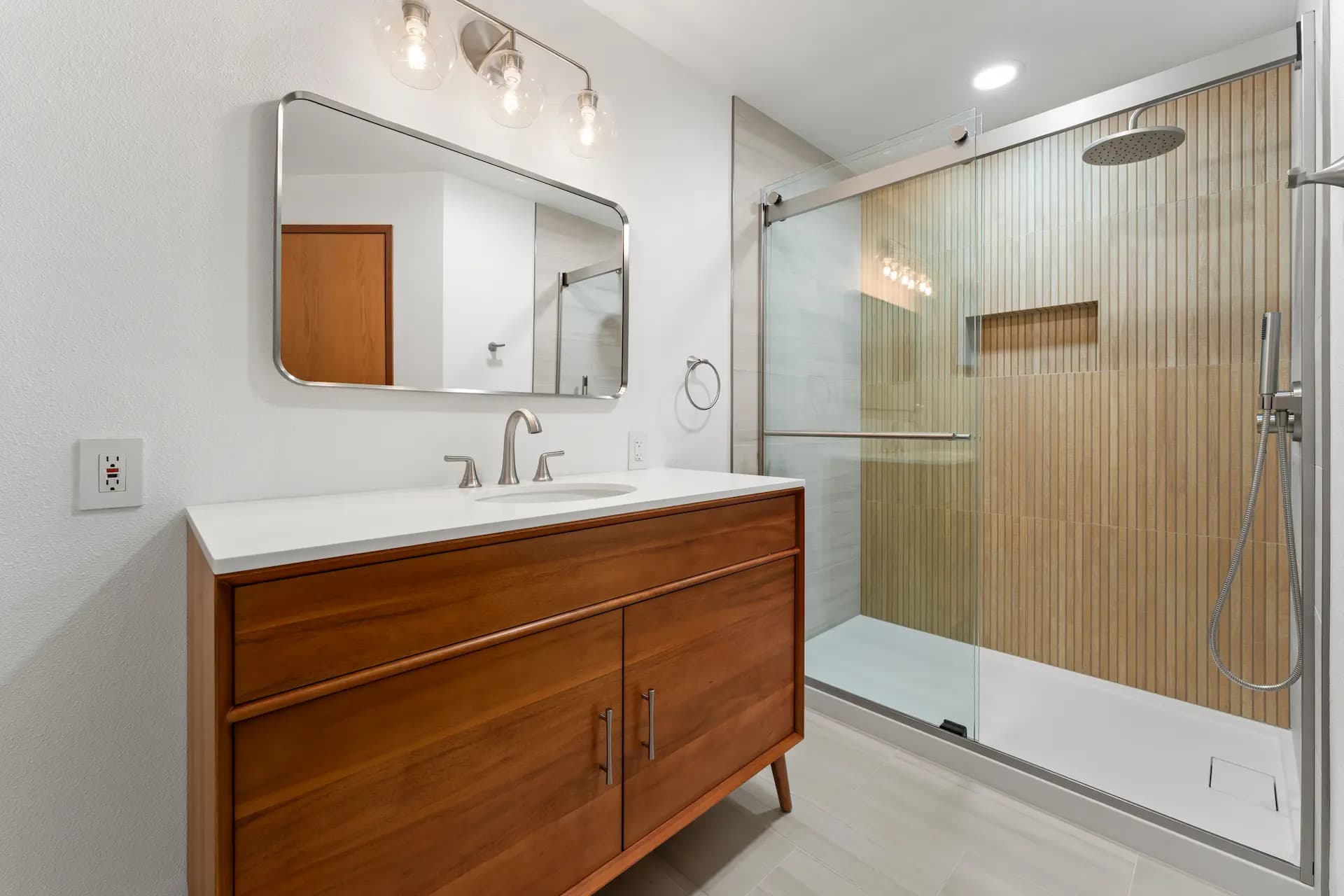 Modern bathroom with wooden vanity, white countertop, large mirror, and glass-enclosed shower with vertical wood panel tiles.