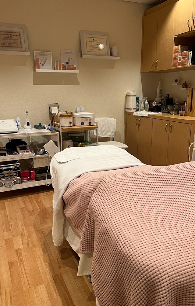 Treatment room with a massage table covered by white and pink blankets, wooden floor, shelves with certificates and skincare products, and wooden cabinets.