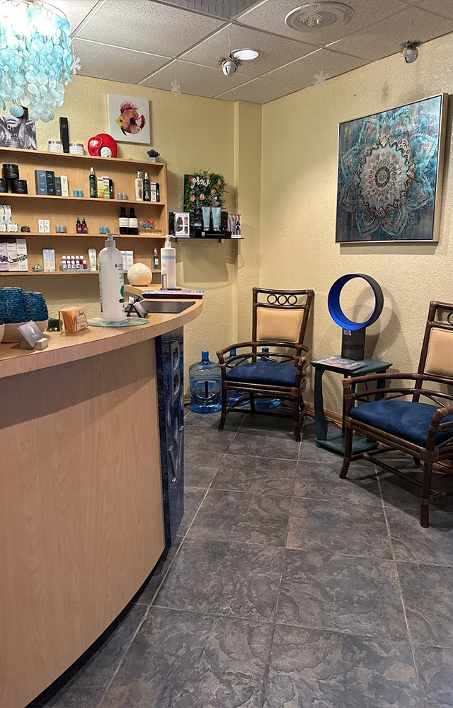 Reception area with wooden counter, shelves holding skincare products, two chairs with blue cushions, a small table with a bladeless fan, and a decorative mandala painting on the wall.