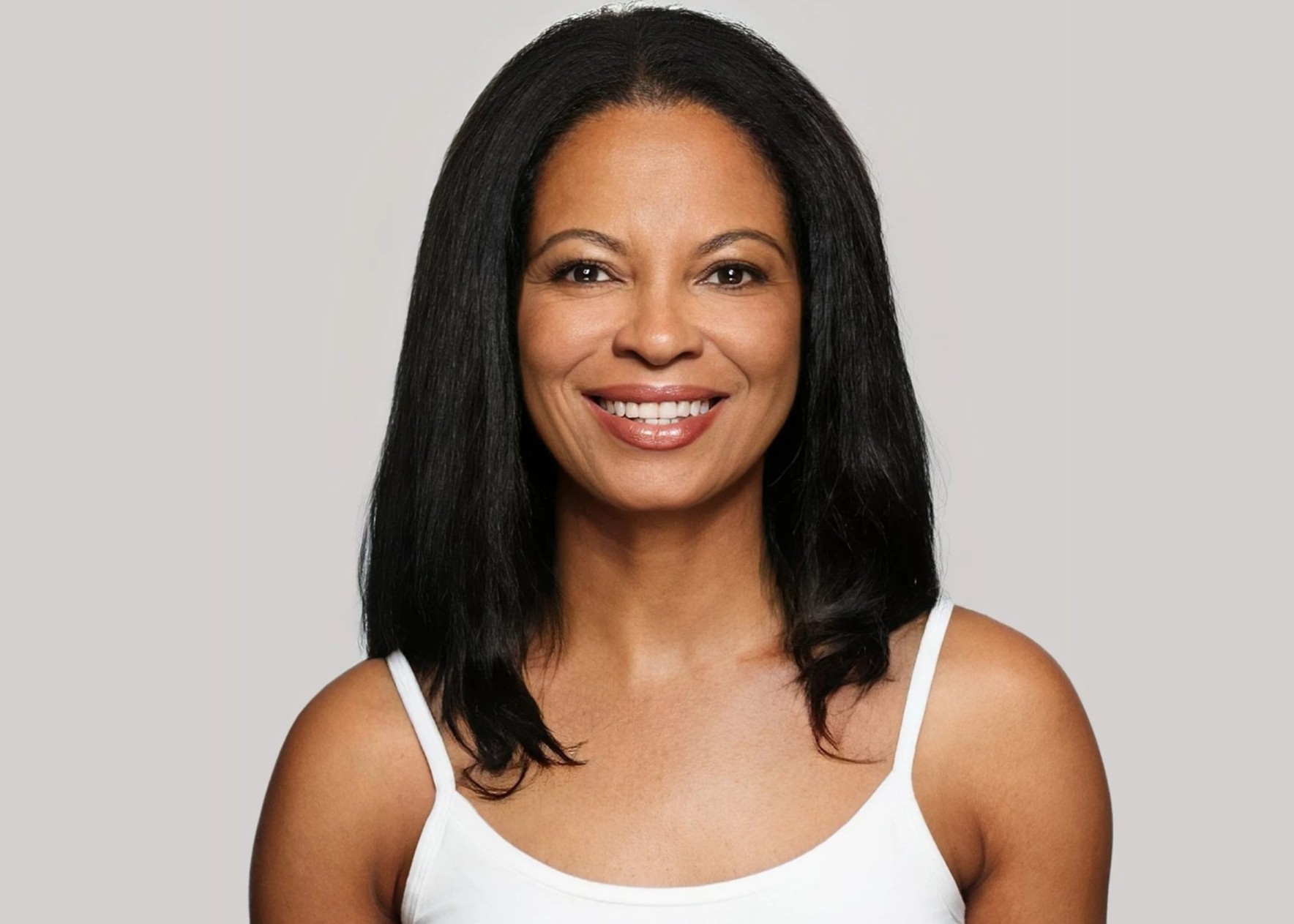 Smiling woman with straight black hair wearing a white tank top against a light gray background.