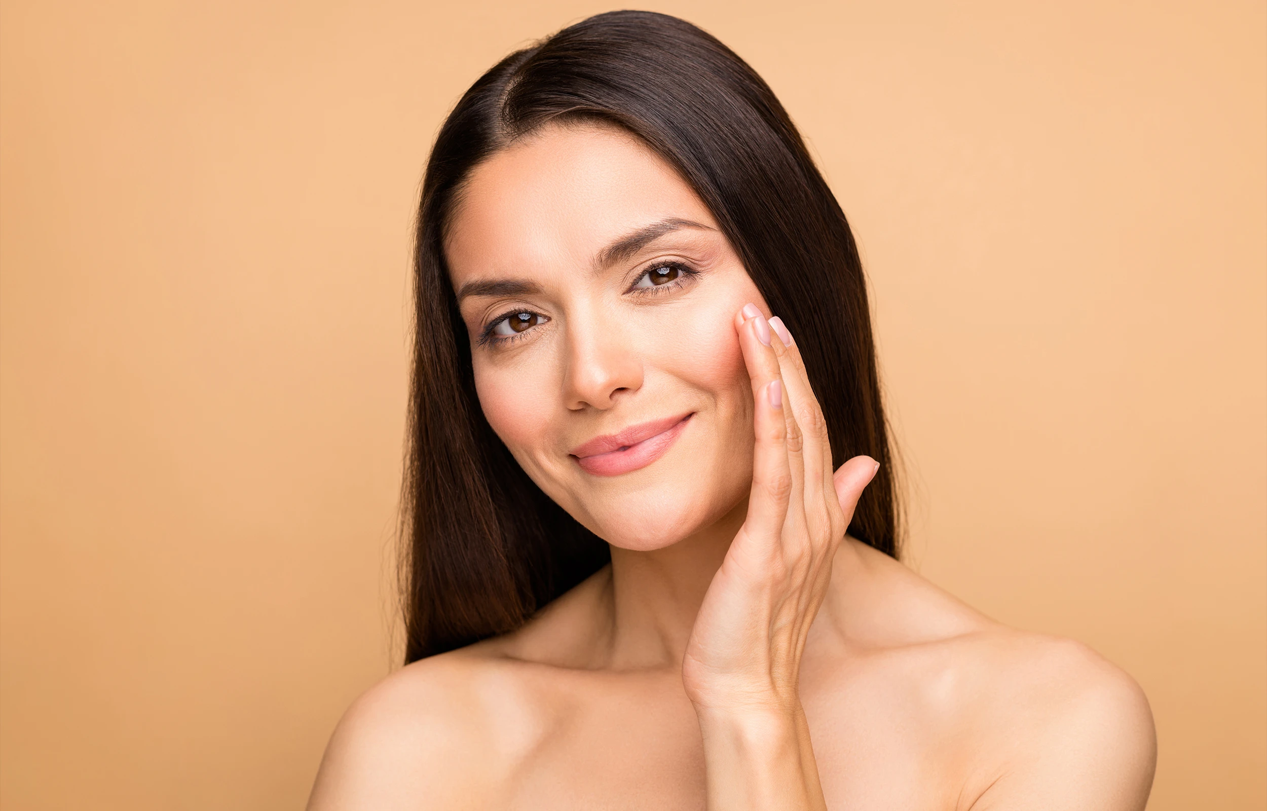 Smiling woman with clear skin gently touching her cheek against a beige background.
