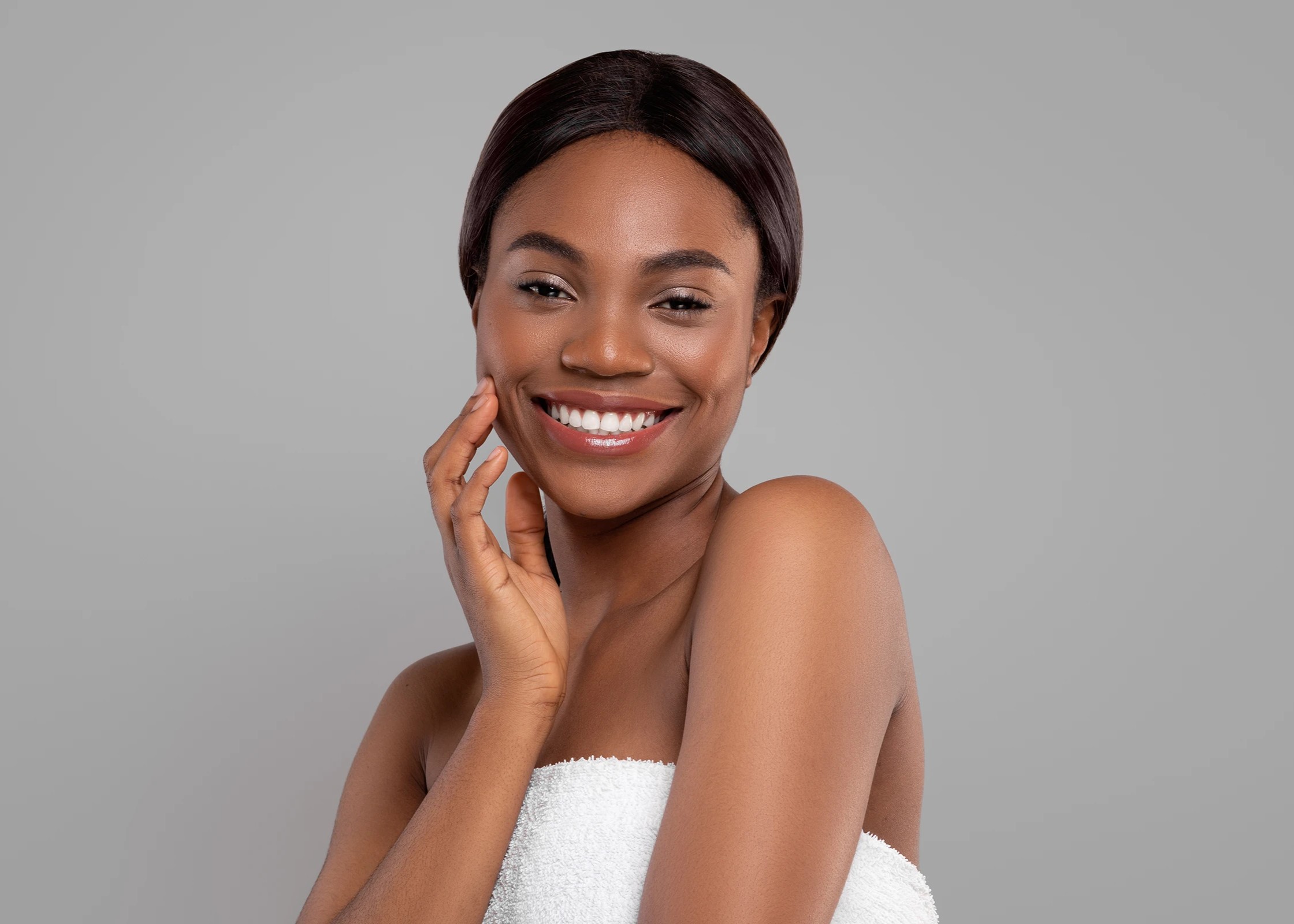 Smiling woman with natural curly hair applying cream to her cheek against a gray background.