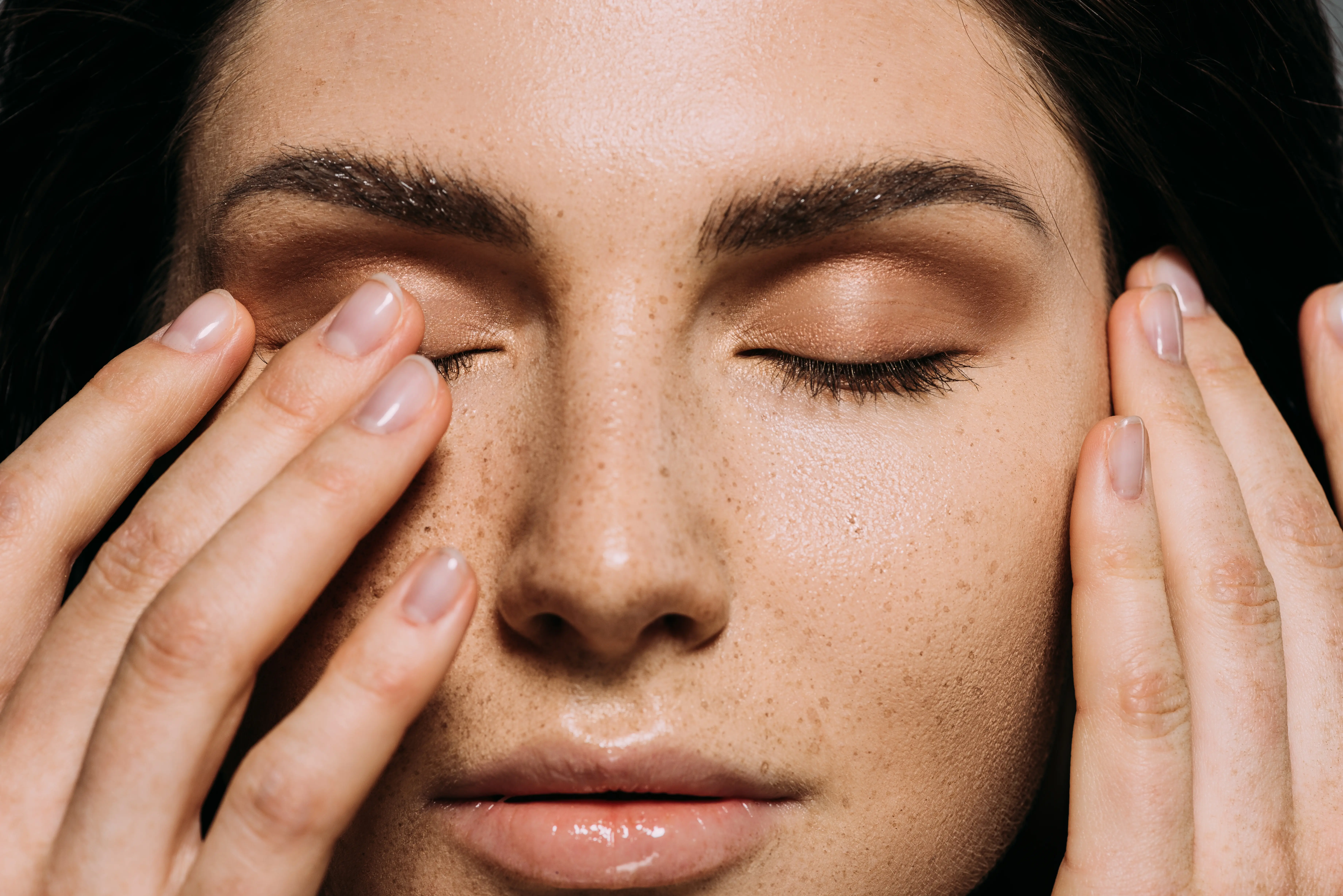 Close-up of a woman's face with eyes closed and hands gently touching her cheeks, showing smooth skin with freckles and subtle makeup.