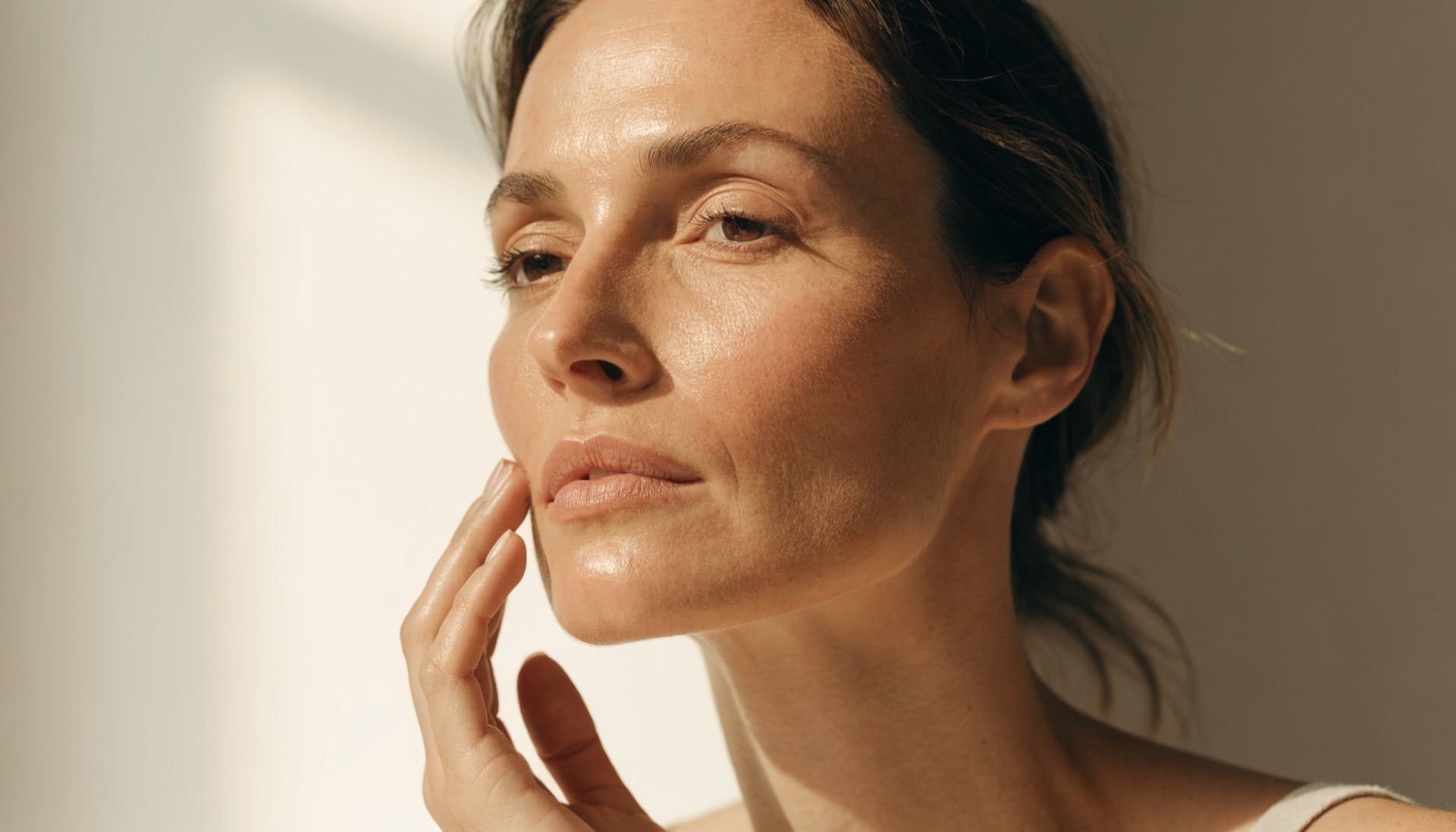 Woman in her forties with natural glowing skin resting her fingertips along her cheekbone and jawline in soft window light