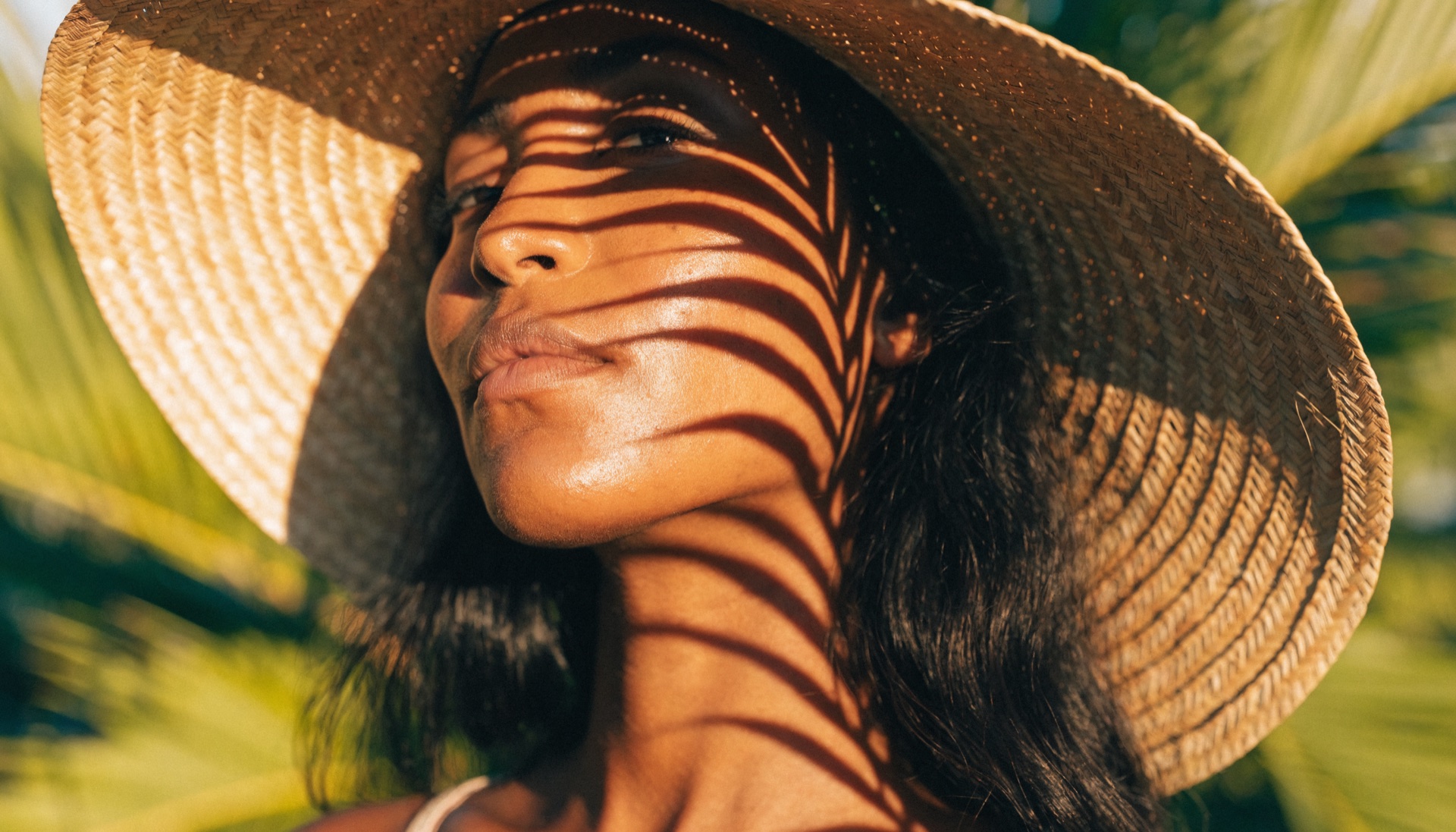 Woman with warm medium deep skin tone wearing a straw sun hat with palm frond shadows across her face in bright Florida sunlight