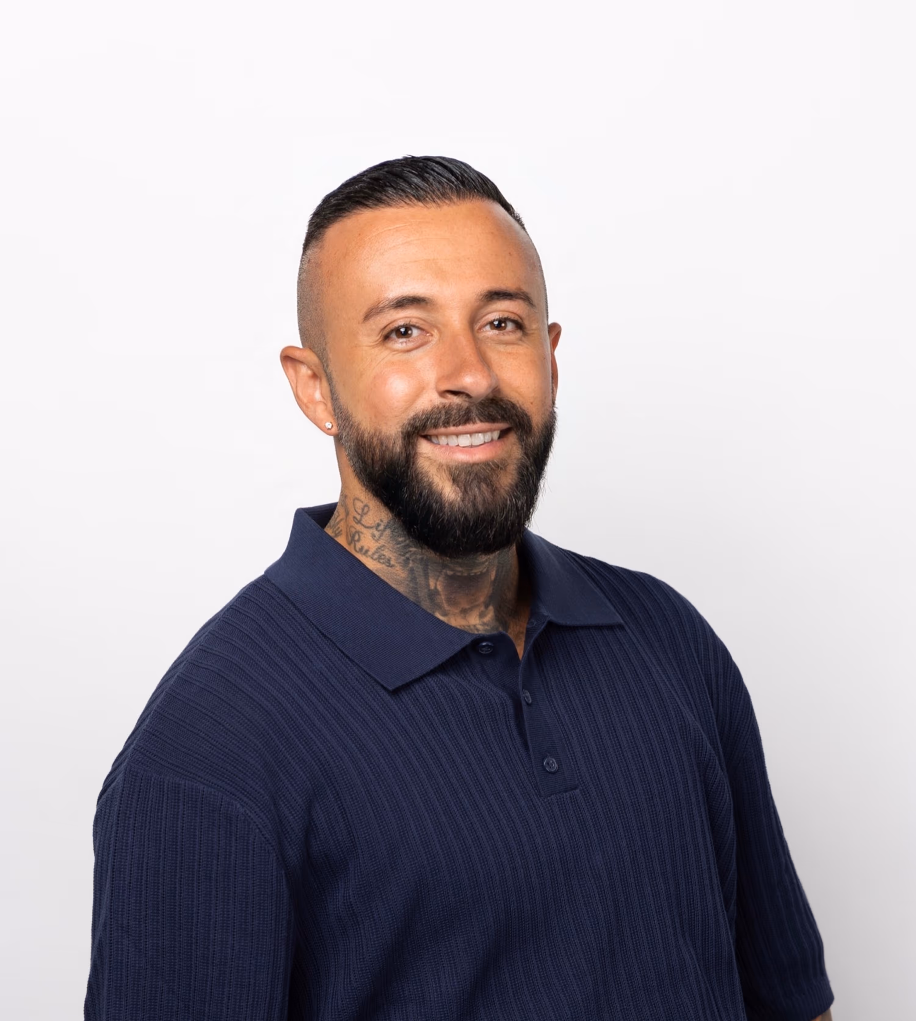 Mann mit kurzem schwarzem Haar, Bart, Hals-Tattoo und blauem, kurzärmligem Pullover lächelt vor weißem Hintergrund.